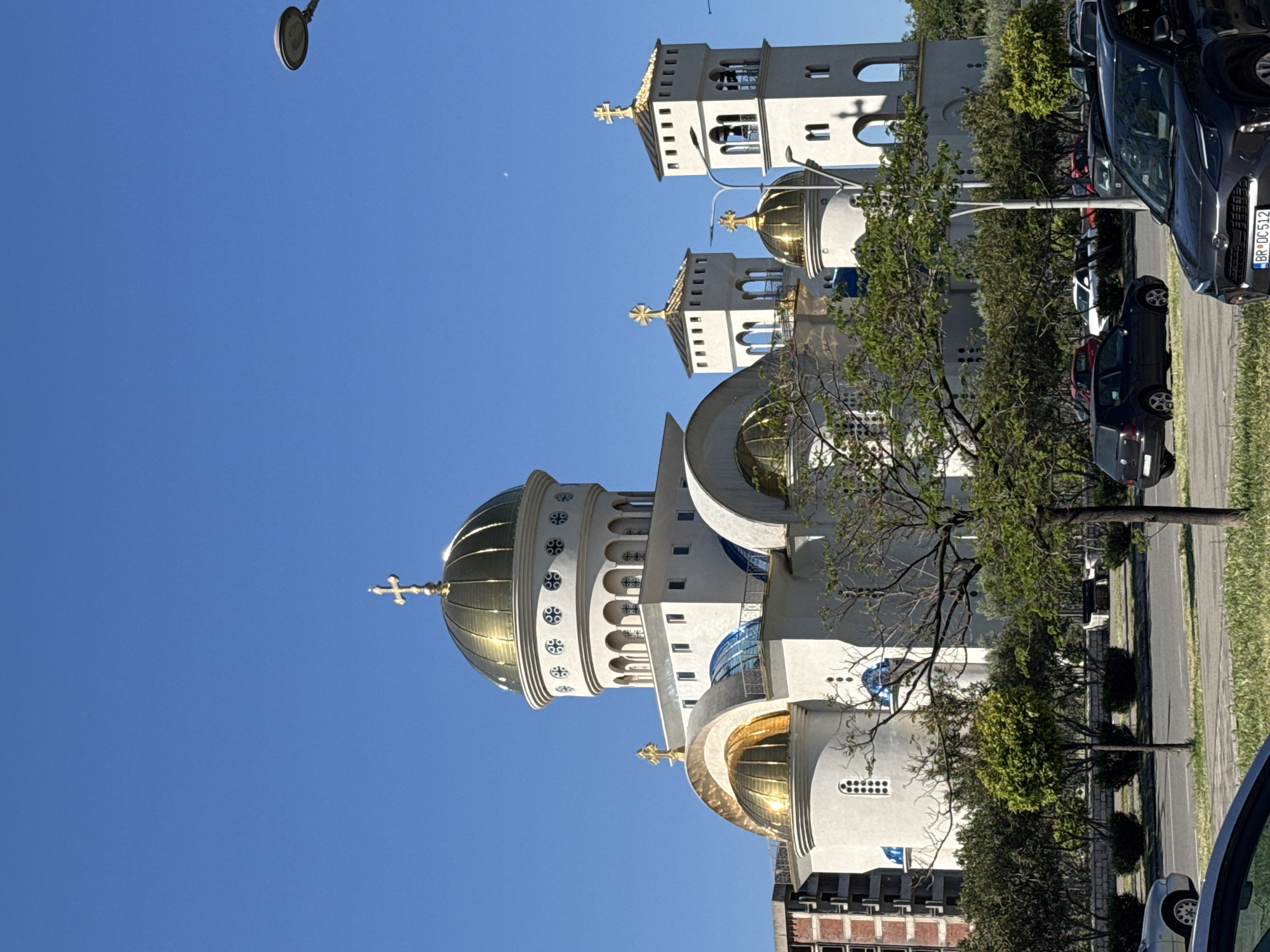 The image depicts the Serbian Orthodox Cathedral of St. John Vladimir in Bar, Montenegro. The building's white exterior and golden domes are visible against a clear blue sky. The cathedral is flanked by trees and a road with parked cars. A street lamp is seen at the top left of the frame. A modern brick and concrete building is partially visible behind the church.