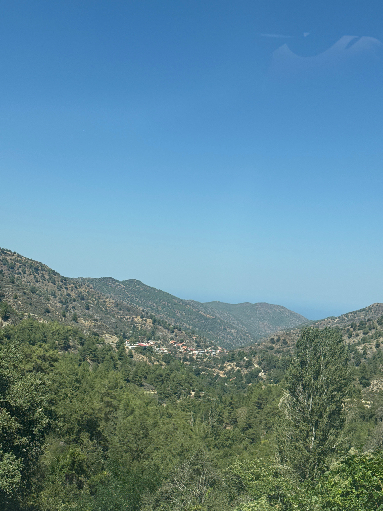 The image depicts a clear, bright day with a vast expanse of blue sky dominating the upper portion. Below the sky, a range of rolling hills covered in sparse, dry vegetation and scattered trees stretches across the landscape. In the mid-ground, nestled within a valley, is a small village characterized by its red-tiled roofs. The village appears to be situated on a gentle slope, with buildings clustered together. The foreground is filled with dense, green foliage, primarily trees with varying shades of green, suggesting a lush, natural environment. The overall scene conveys a sense of a rural, possibly mountainous, setting in Pedoulas, Cyprus.