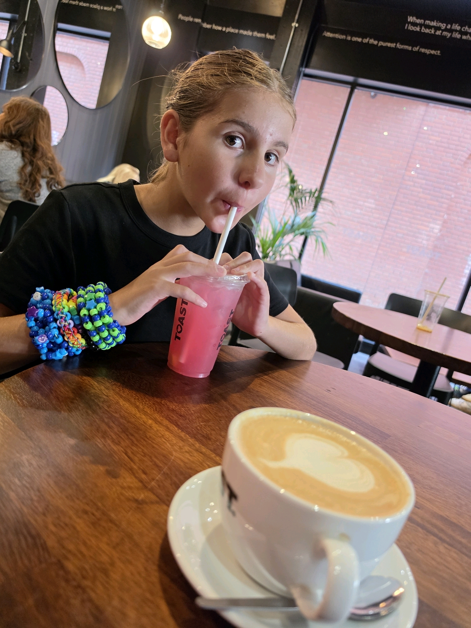A young person is seated at a wooden table in a cafe, drinking a pink beverage through a straw from a plastic cup. They are wearing a black t-shirt and have several colorful, beaded bracelets on their left wrist. In the foreground, a white cup with a latte art design of a heart sits on a saucer with a spoon. The cafe has large windows with a view of brickwork and blinds, and mirrors are visible on the wall in the background. Text is present on the walls above the windows.