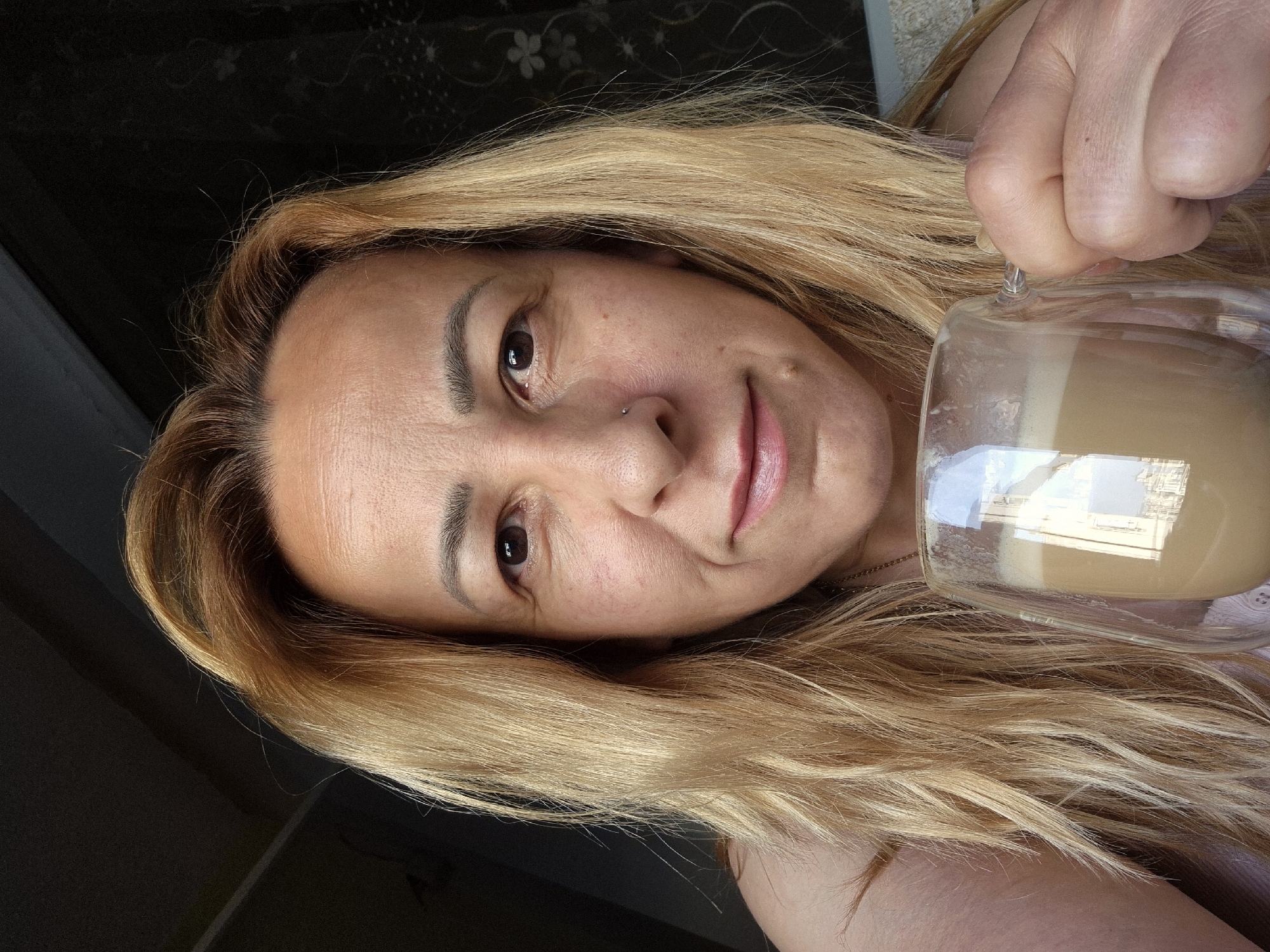 A woman with long, blonde hair holds a glass mug containing a light brown beverage. She has light skin, brown eyes, and a nose piercing. The background is blurry, but a dark, floral patterned surface is visible. The location is Unknown, Unknown.