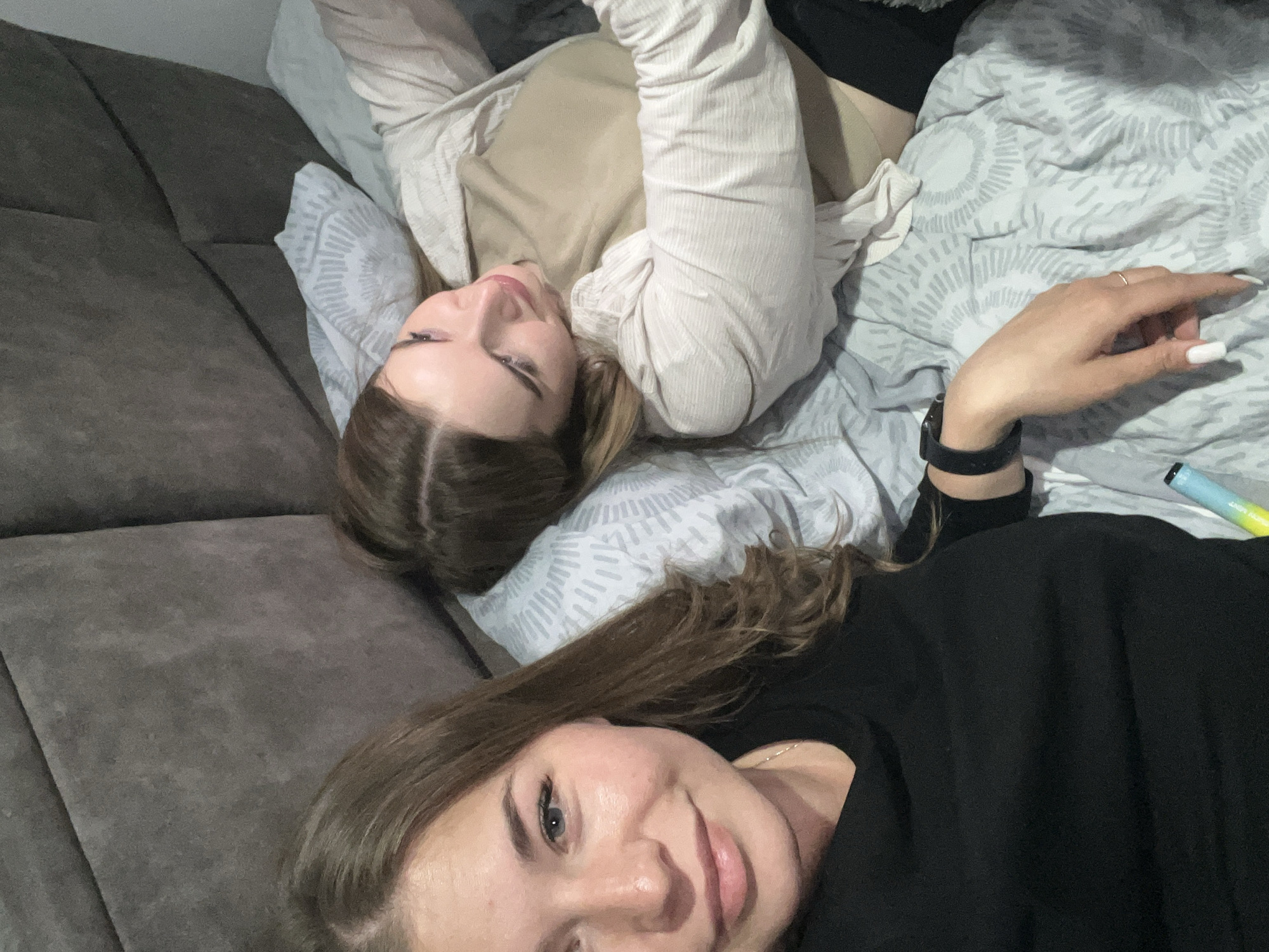 The scene depicts two adult females indoors, likely within a private residence or hotel room in Budva, Montenegro. Both individuals are positioned on a bed with a dark grey upholstered headboard and light blue-grey patterned bedding.
The female closer to the camera, on the left side of the frame, has long, light brown hair and is looking directly at the viewer with a slight smile. She is wearing a black long-sleeved top. Her left hand, visible in the foreground, features a black smartwatch on her wrist and a simple ring on her ring finger, with her fingernails manicured with white tips. A light blue and green disposable vape pen rests on the bedding near her left hip.
The second female, positioned further back on the right, is lying on her back with her head resting on a pillow. She has brown hair and is looking towards the left, in the direction of the first female or the camera. She is wearing a light beige long-sleeved shirt over a tan undergarment, with black fabric, possibly part of trousers or shorts, visible beneath the bedding. The background reveals a plain white wall to the right of the bed.