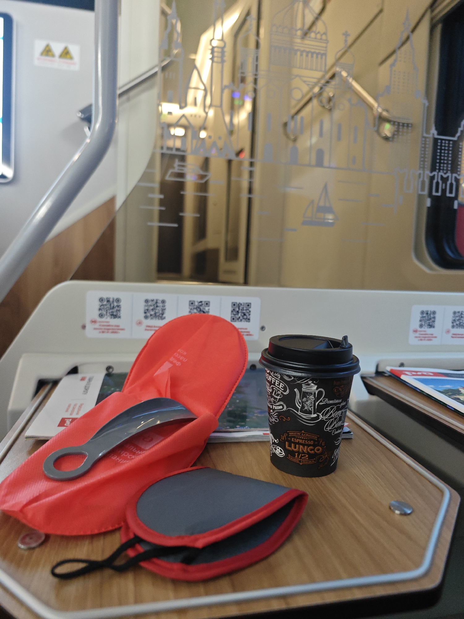 This image depicts a tray table on a train, likely in Saint Petersburg, Russia, given the distinctive architectural decorations on the train's interior. The tray table is made of wood and has a metallic edge. On the tray, there is a black disposable coffee cup with "COFFEE" written in decorative lettering and "LUNCO" printed on it. Next to the cup, there is a folded red fabric pouch with a gray shoehorn inside. To the right of the pouch, there is another folded item, possibly a case, with red trim and a gray exterior. Behind these items, on the tray, are some papers, including a magazine or brochure with an image of a landscape. On the wall behind the tray table, there are several QR codes. The background shows the interior of the train carriage, with glass panels decorated with stylized drawings of St. Petersburg landmarks, such as cathedrals and a sailboat. The lighting suggests an indoor environment.