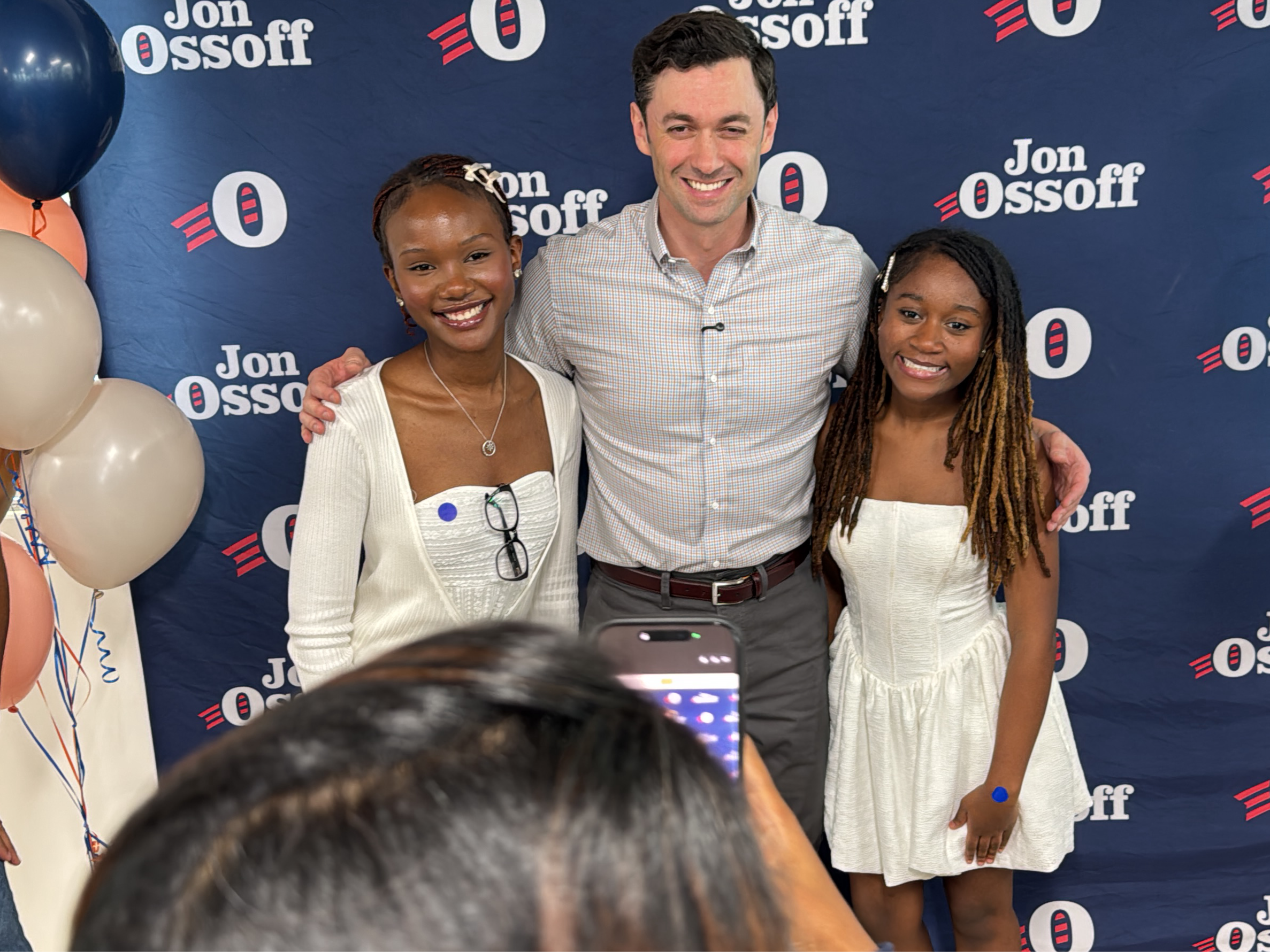 The image shows a group photo of three people standing in front of a backdrop with the name "Jon Ossoff" repeated. The central figure is a light-skinned man wearing a button-down shirt and gray pants. To his left is a dark-skinned young woman in a white top and cardigan, and to his right, another dark-skinned young woman is wearing a white dress. All three are smiling. A hand holding a cell phone is visible in the foreground, suggesting someone is taking the picture. Balloons are present on the left side of the image. The location is the city of Decatur, United States.