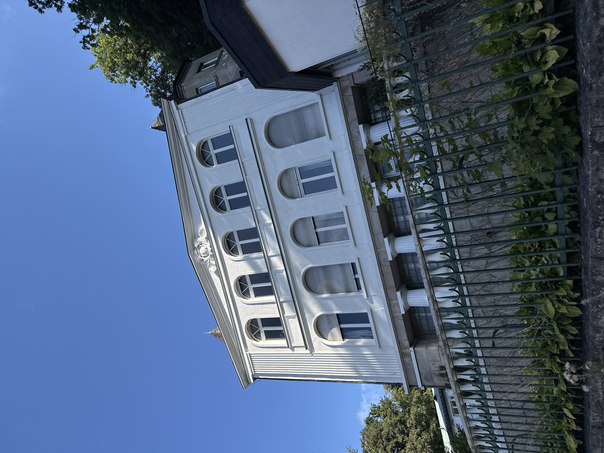 The image shows a white building with classical architectural elements, including arched windows and columns, under a clear blue sky. The building is multi-storied and appears to be residential or a public institution. It is situated on a slope, with a green metal fence in the foreground bordering a stone wall. Lush green foliage grows along the fence and behind it, partially obscuring the lower level of the building. Trees are visible on the right and left sides of the building, suggesting a natural or park-like setting. This building is identified as the "Haus am Park" in the city of Bad Sachsa, Germany, a well-known spa town.