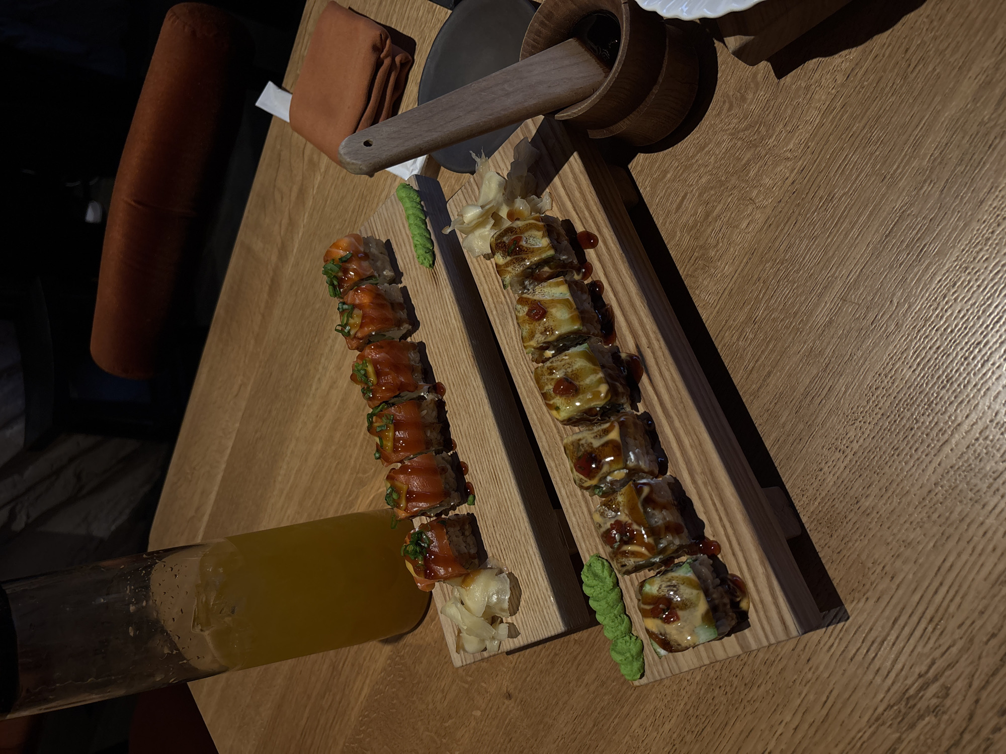 A high-angle shot depicts a wooden table setting with sushi, soy sauce, wasabi, and a glass of juice. The sushi is arranged on a wooden serving board, consisting of two rows of different types. Adjacent to the sushi are mounds of wasabi and pickled ginger. A dark wooden container with a handle, presumably holding soy sauce, is positioned near the top of the sushi board. A glass of yellow juice sits beside the left row of sushi rolls. The table has a light brown, wood-grain finish. Part of a brown upholstered chair back is visible on the left. The location may be a restaurant in Krasnoyarsk, Russia, based on visual elements of the image, although this cannot be confirmed.