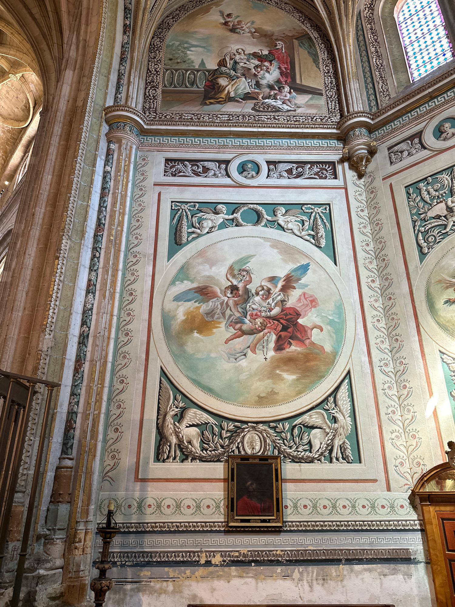 The image depicts a section of the interior of a historical building, likely a church or a monastery, located in La Lastrilla, Spain. The scene is dominated by ornate wall decorations and a large fresco.
On the upper part of the wall, a fresco depicts several figures in a scene that appears to be religious in nature, possibly a biblical narrative. Below this, a large circular fresco is the central focus. This fresco features cherubic figures with wings, often referred to as putti or angels, gathered around a central element. One angel, dressed in red, holds a baby or young child who is wearing a rosary. Other winged figures are also present, with vibrant colors and delicate brushwork.
Surrounding the circular fresco are decorative borders and smaller framed artworks or medallions. These include griffins or mythical winged creatures depicted in relief or painted on the wall, surrounded by wreaths of leaves. A smaller framed painting, hanging below the circular fresco, shows a dark interior with a figure in red.
The architectural elements visible include stone pillars and arched structures, hinting at a grand and ancient construction. The walls themselves are adorned with intricate patterns, including geometric motifs and floral designs, executed in various colors like green, red, and gold. The overall impression is one of rich artistic detail and historical significance. The lower part of the wall shows signs of wear and tear, with peeling paint and plaster, suggesting the age of the structure. A wooden candle holder with a single candle is visible on the left side of the image, further adding to the historical atmosphere. A stained-glass window with a diamond pattern is visible in the upper right corner.