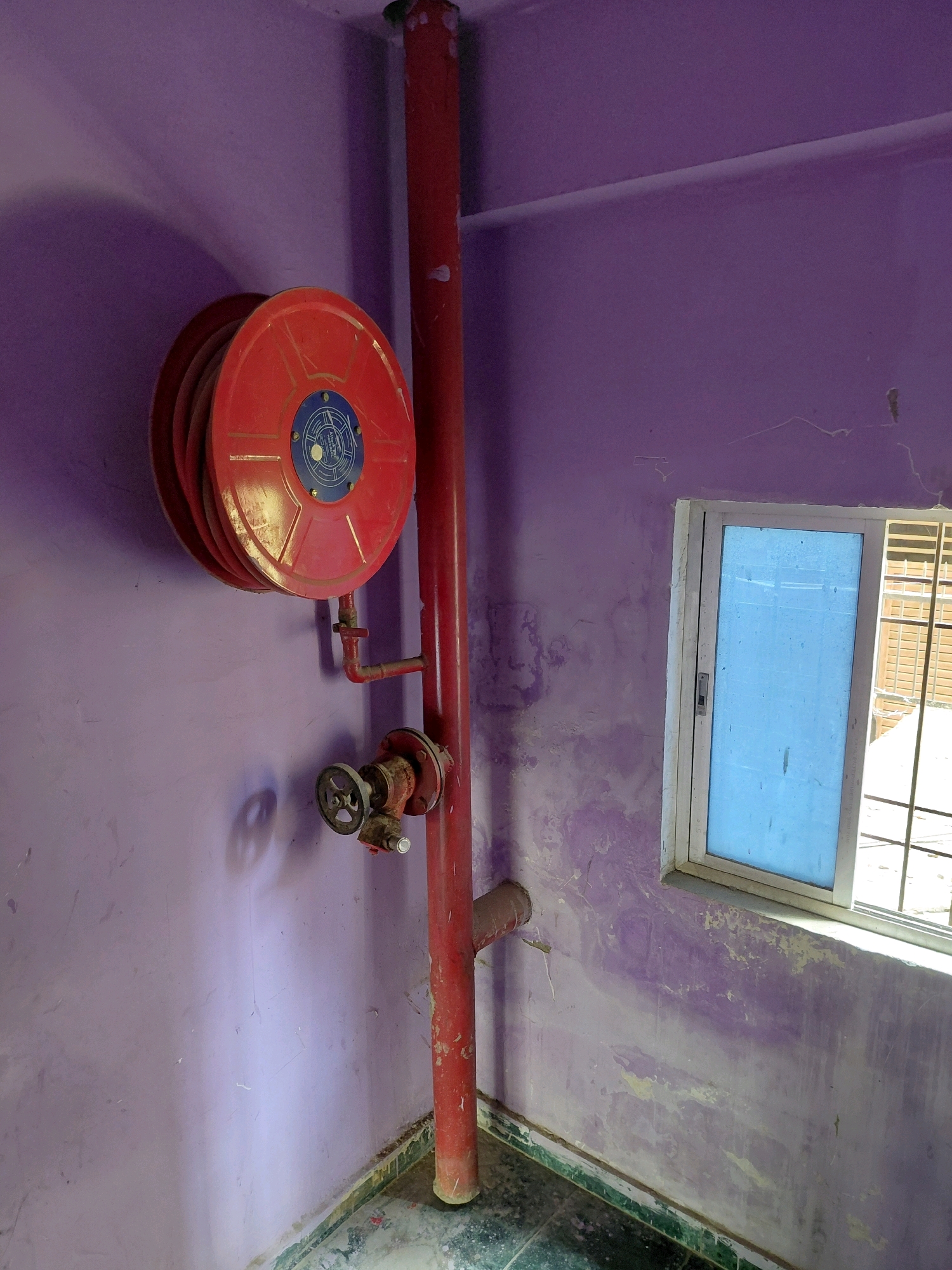 The image depicts a fire hose reel mounted on a purple wall in an interior space. A red pipe runs vertically along the wall, connected to the hose reel and a valve. To the right of the fire safety equipment is a window with a light blue pane, covered by a grate on the exterior. The walls show signs of peeling paint and water damage, particularly near the floor where a green border is visible. The floor appears to be tiled. This scene is likely located in Patna, India, given the context. There are no people or animals visible in the image, and no actions or interactions are taking place.