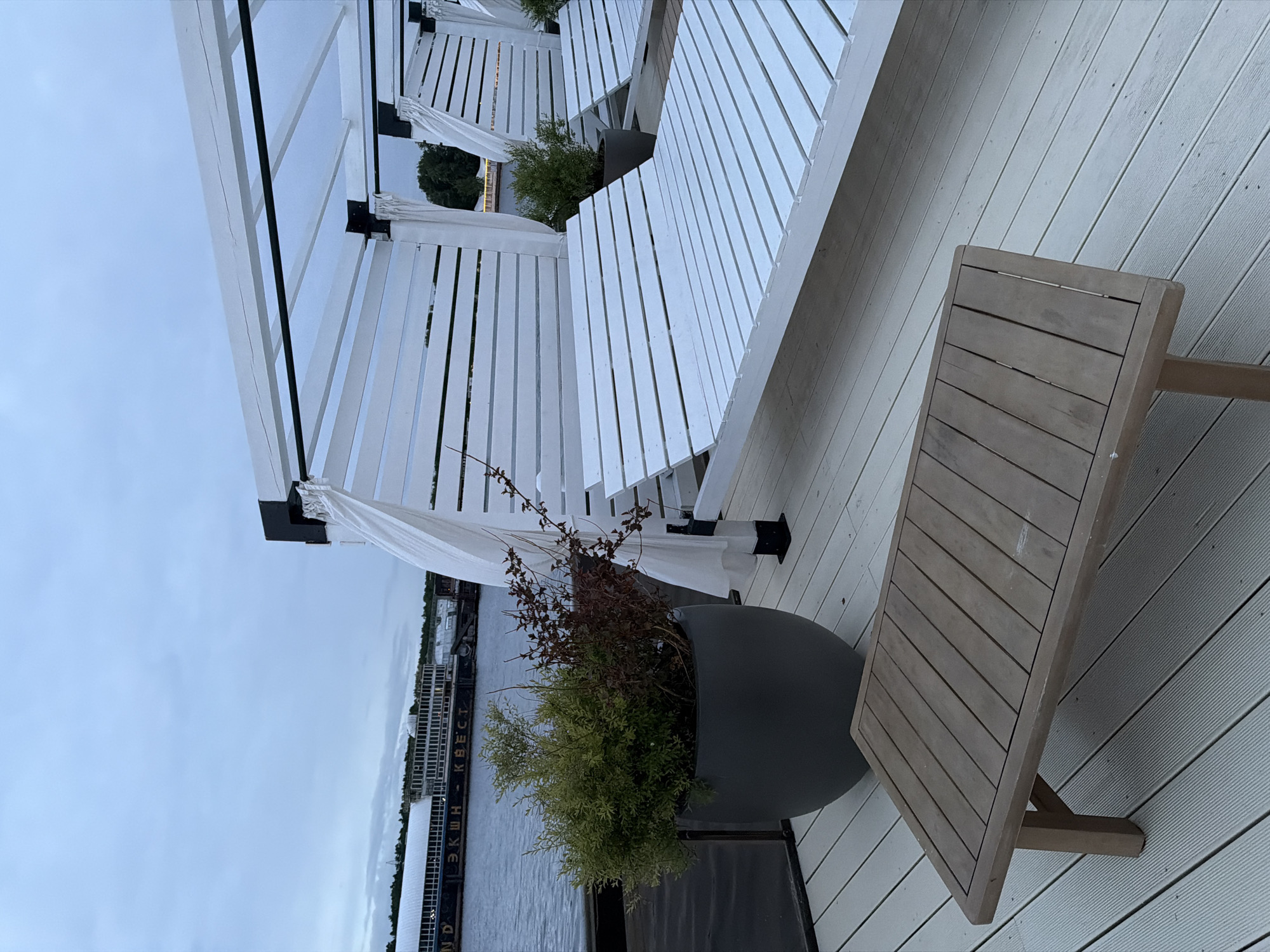 A medium, close-up shot depicts an outdoor patio scene overlooking a body of water in an unidentified city. The patio is equipped with white pergolas draped with white fabric. A gray planter sits in the foreground with a mixed collection of green shrubs and bare brown branches. A wooden table stands next to the planter. In the background, a long, dark-colored ship-like structure is visible on the water. The sky appears overcast.