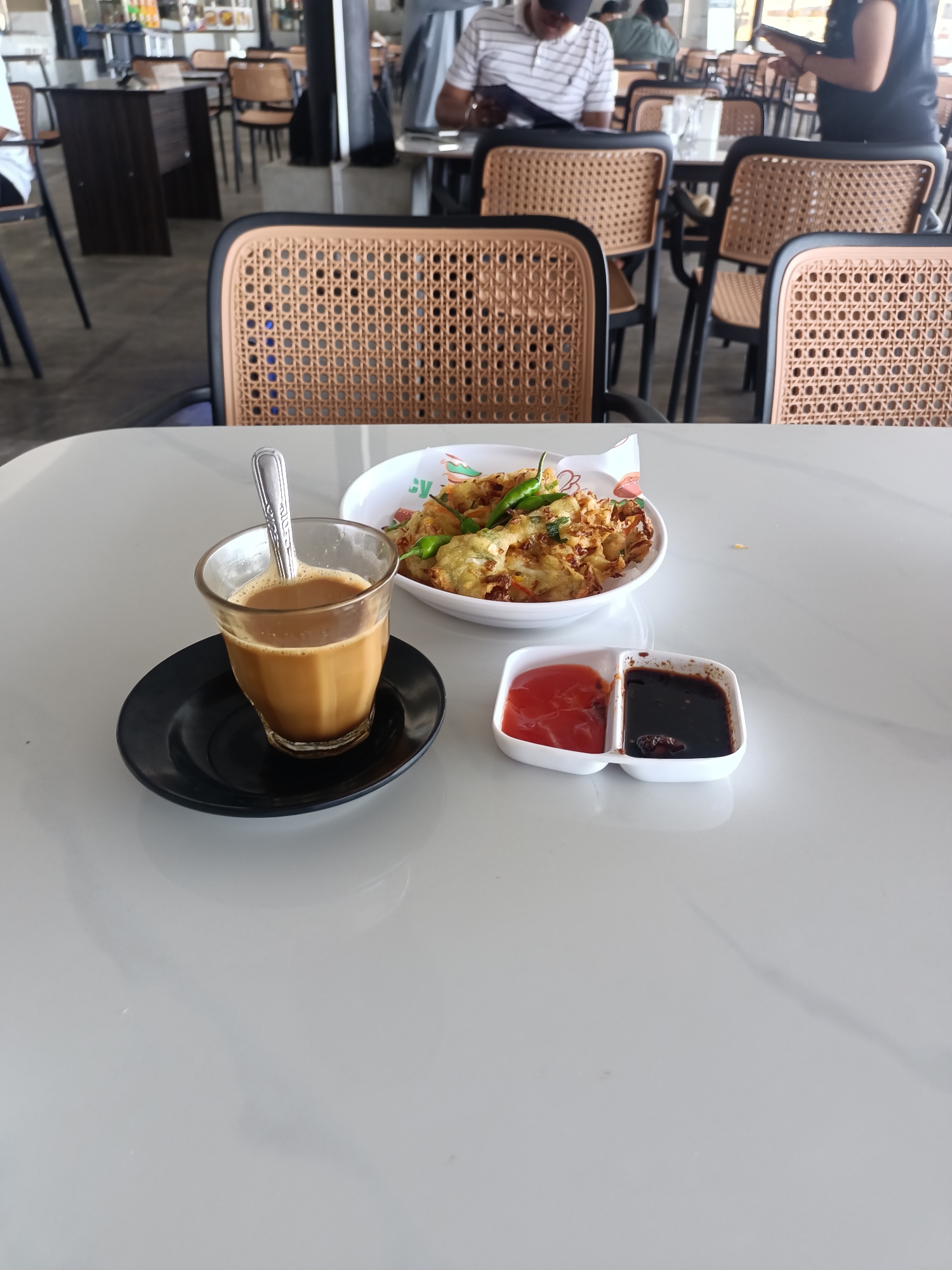 The image presents a candid shot of a meal set on a white, glossy table within a cafe or restaurant in Batam, Indonesia. In the foreground, a clear glass containing a light brown beverage, likely coffee or tea with milk, stands on a black saucer, with a silver spoon resting in the drink. Adjacent to it, a white bowl holds several fried fritters, such as bakwan, topped with fresh green chilies. A small, white, two-compartment dish contains a vibrant red sauce (possibly chili sauce or ketchup) and a dark brown sauce (likely soy sauce or sweet soy sauce) for dipping. The paper liner beneath the fritters shows partially visible text "SY" and "B," along with a colorful, fruit-themed border.
The background, intentionally blurred, depicts a busy cafe setting. Numerous modern chairs with black frames and woven rattan-patterned backs are arranged around dark tables. On the left, a man in a striped shirt and dark cap is seated, looking down, possibly at a menu or phone. On the right, another individual in a dark shirt stands, holding what appears to be a tablet or menu. Other blurry figures are visible throughout the cafe, indicating a daytime setting. The floor appears to be a grey, industrial-style concrete. The overall scene captures a casual dining or snack experience.