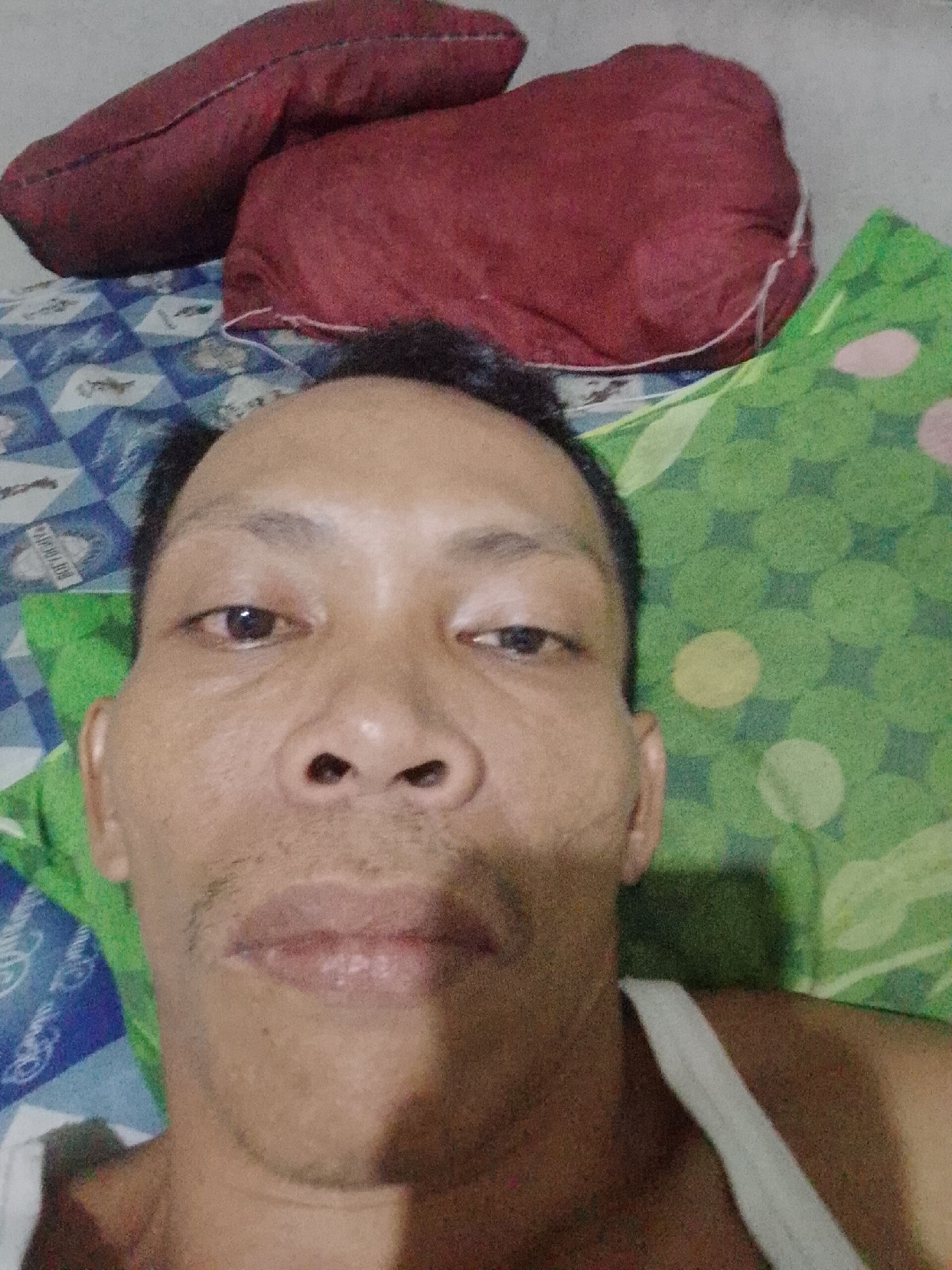 A man with short black hair and tan skin lies on a bed with a blue and white patterned sheet and a green pillow. Two maroon pillows rest on the sheet near his head. He wears a white tank top. The photo was taken in the city of Bukit Ketok, Indonesia.