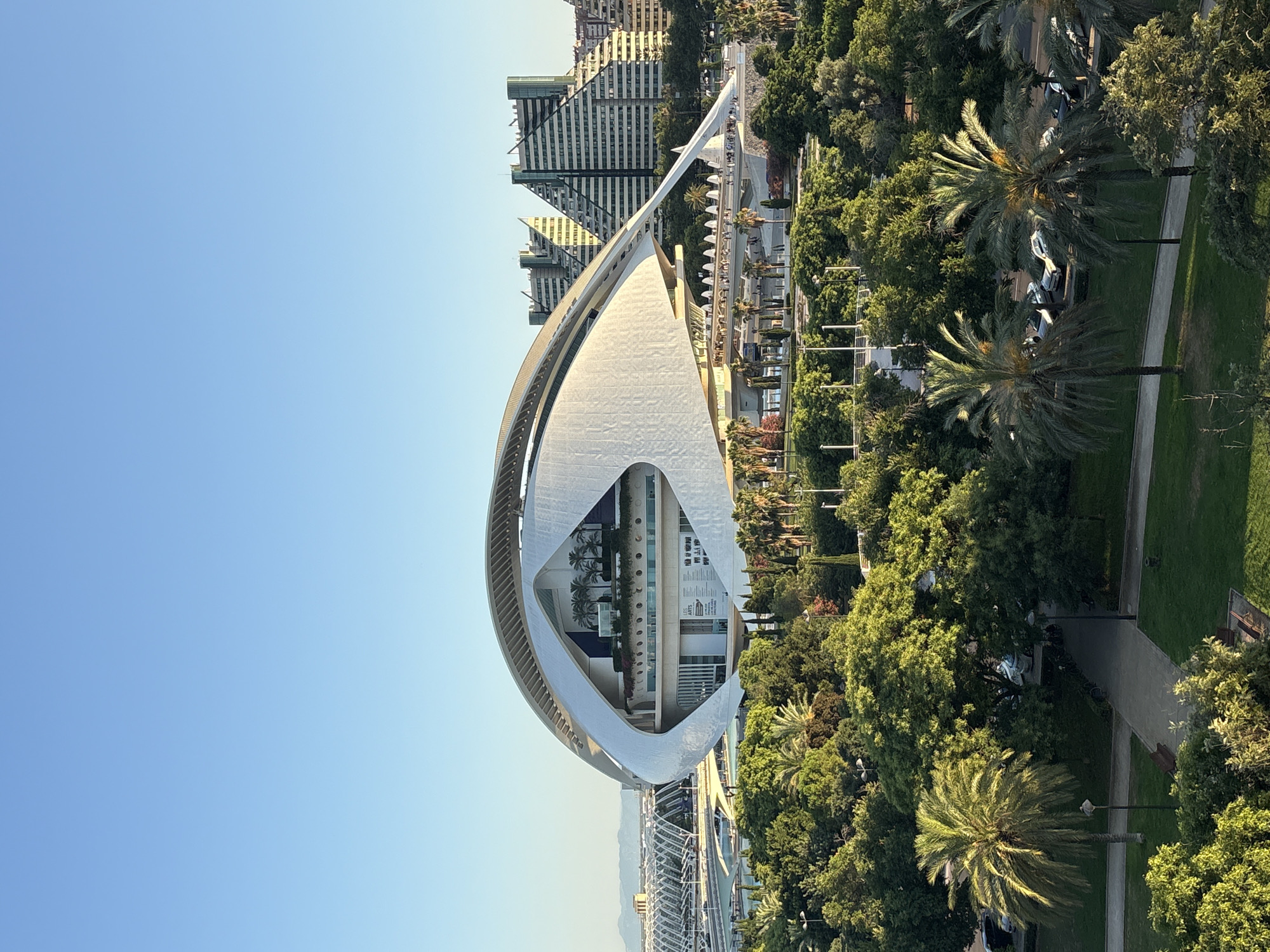 The image shows a clear, bright day with a wide expanse of pale blue sky dominating the upper portion. In the mid-ground, a large, distinctive modern building with a white, curved, architectural design is prominently featured. This building appears to be part of a complex, possibly a cultural or performance venue, with multiple levels visible and greenery, including palm trees, on a raised platform.
Below and in front of this main structure, there is a lush green park area with numerous trees, including several palm trees. The park features pathways, benches, and lampposts, suggesting a public space. In the distance, behind the main modern building, other buildings with angular, stepped architectural designs are visible, indicating an urban setting.
Further in the background, what appears to be a bridge or elevated walkway structure with an arched design can be seen, along with more distant buildings and a hazy outline of mountains on the horizon. Several cars are parked in an area adjacent to the park. The overall scene is one of modern architecture integrated with green spaces within a city environment. This building is the Palau de les Arts Reina Sofía, located in the City of Arts and Sciences in Valencia, Spain.
