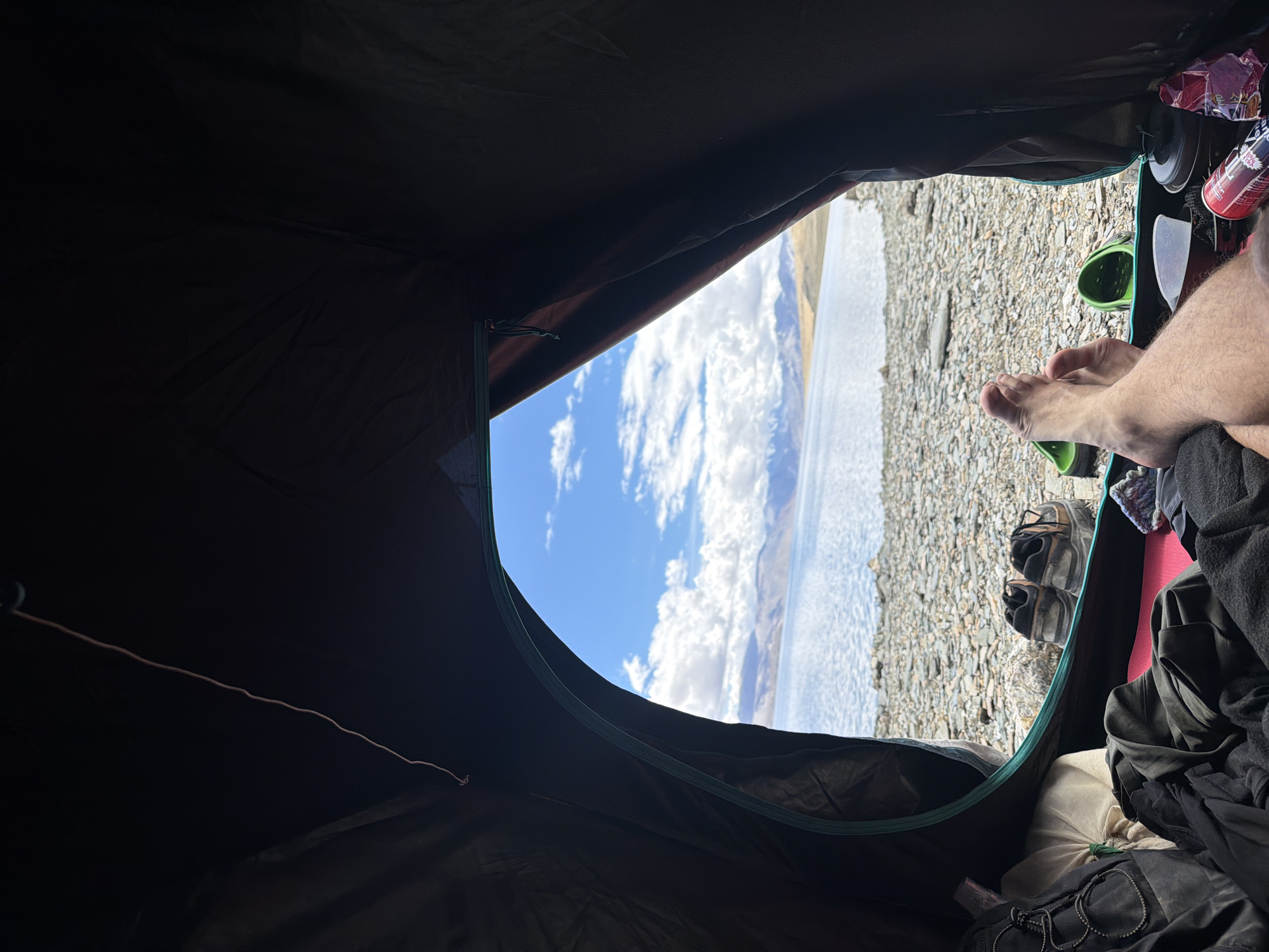 An interior view from inside a tent shows a person's bare legs and feet resting on a sleeping bag, looking out at a tranquil lake, rocky shore, and distant mountains under a partly cloudy sky. Two pairs of shoes are visible near the tent entrance, one pair of hiking boots and a pair of dark casual shoes. Several items of camping gear, including a backpack, clothing, a metal mug, and a can of food, are scattered inside the tent. The scene suggests a peaceful camping experience in a remote, natural setting.