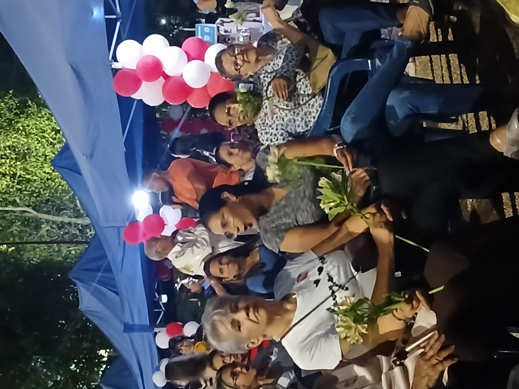 The image shows a gathering of people seated outdoors in Ciudad Guayana, Venezuela. A blue tent covers part of the scene, and red and white balloons are visible. People are holding flowers. Some are wearing casual clothing like jeans and patterned shirts. Chairs are visible, and the environment appears to be a public space.