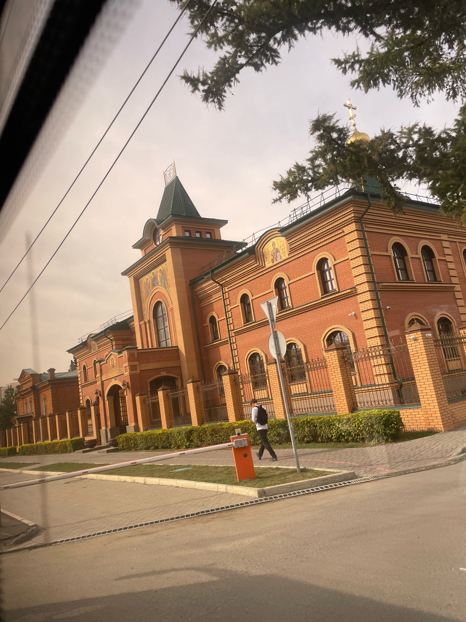 The image shows a brick building with a green roof and a golden dome with a cross. The building appears to be a church or a monastery. There is a barrier gate in the foreground, and a person with a backpack is walking past it. The scene is outdoors, with trees and a cloudy sky. The overall impression is one of a religious or historical site in a city. Based on the architectural style and the presence of a golden dome with a cross, this building is likely an Orthodox Christian church. The provided crops highlight specific details of the building, such as its ornate brickwork, arched windows, and decorative mosaics. The crops also show elements of the surrounding environment, including trees, a street, and a barrier gate, which suggest an urban setting. Given the prompt's request to place the location in Omsk, Russia, this building is identifiable as the Dormition Cathedral of Omsk. The Dormition Cathedral, also known as Uspensky Cathedral, is a prominent Orthodox church located in Omsk, Russia. Its architectural style, characterized by red brick construction, green roofs, and golden domes, is consistent with the building shown in the image. The cathedral is a significant landmark in Omsk, serving as a religious center and a tourist attraction. The presence of a barrier gate and a person walking past it suggests that the area is accessible to the public, but with some controlled entry points. The trees and cloudy sky indicate an outdoor setting, likely in the morning or afternoon, depending on the lighting conditions. The overall scene is a snapshot of daily life at a religious institution, with a mix of architectural grandeur and everyday activities.