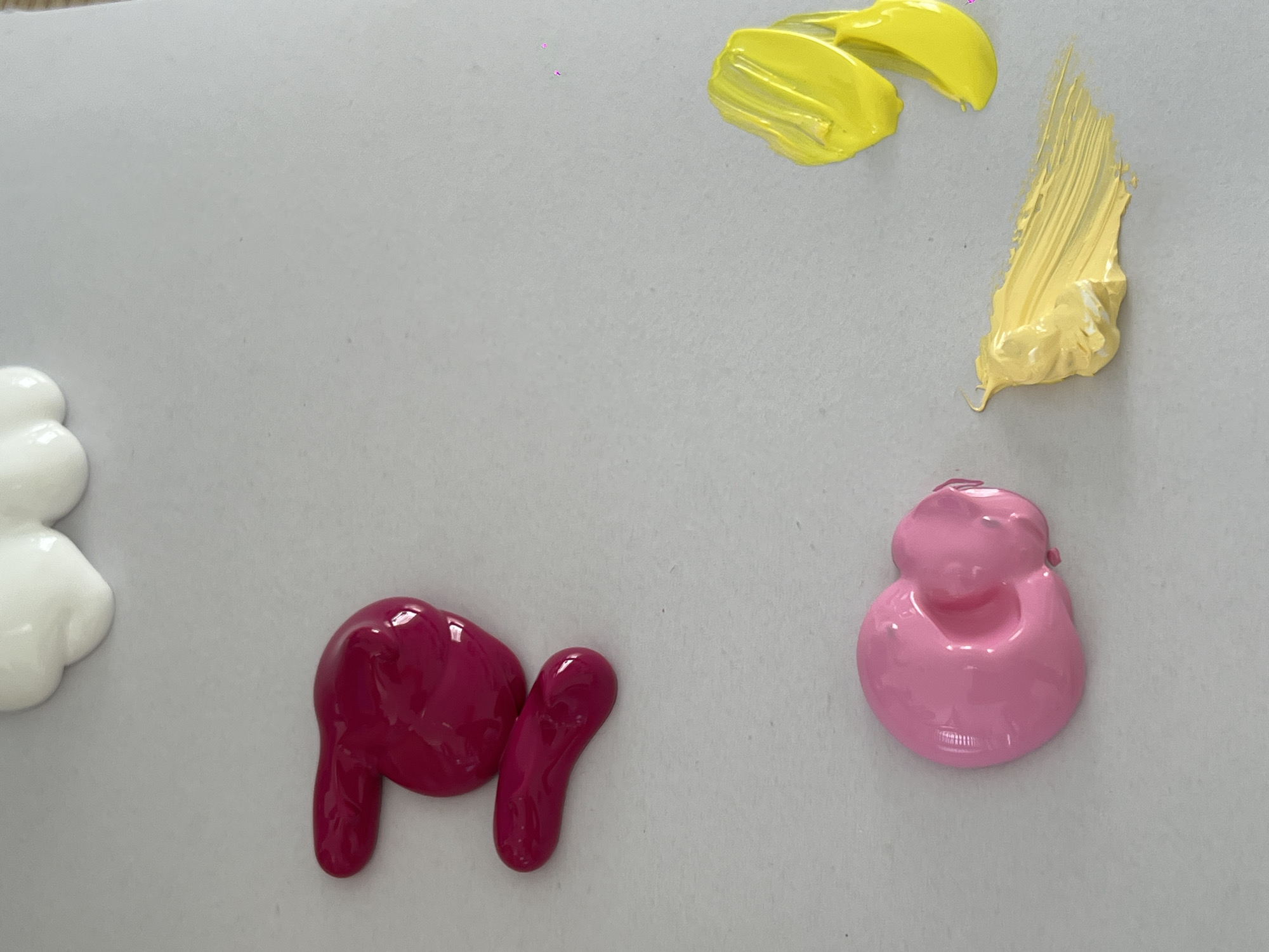The image shows a close-up overhead view of several blobs and brush strokes of paint on a light gray surface. There are two blobs of white paint at the top, one of which is partially visible. Below these are two blobs of bright pink paint. Further down and to the left, there is a blob of light pink paint. To the right and slightly below the pink blob, there are two brush strokes of bright yellow paint, one on top of the other. Below and to the left of these, there is a wider, more textured brush stroke of pale yellow paint. There are no people, city elements, or specific geographical features visible.