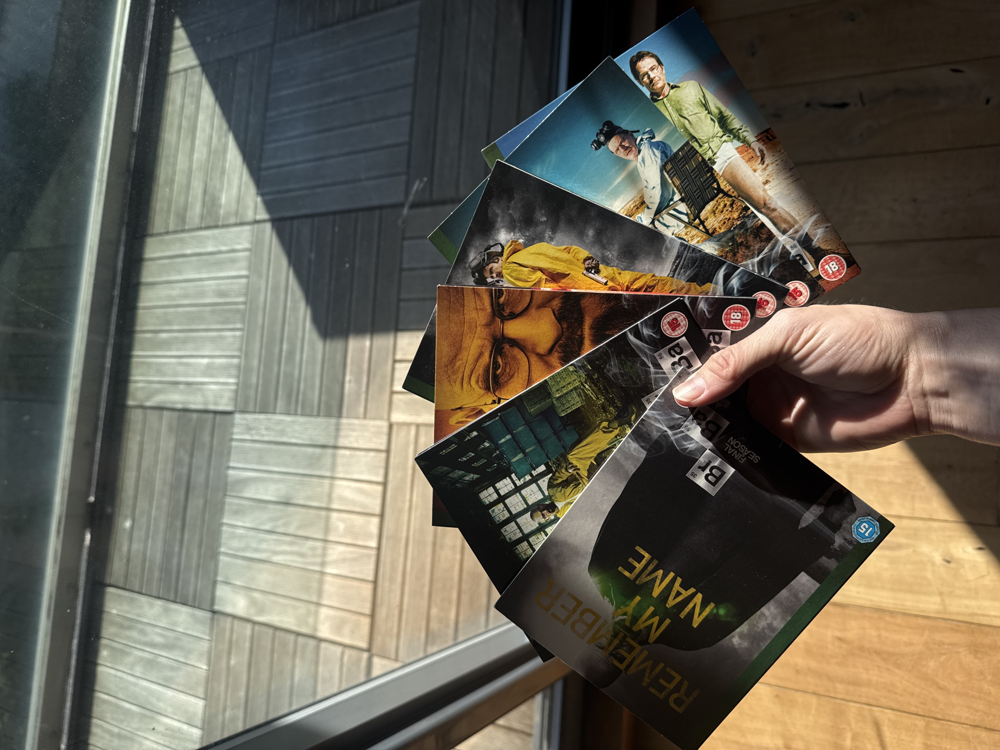 A hand holds a fanned stack of several media cases, likely DVD or Blu-ray discs, in bright sunlight. The cases are predominantly themed around the television series "Breaking Bad."
The topmost visible case features Walter White (Bryan Cranston) in a light green shirt and white shorts, standing alongside Jesse Pinkman (Aaron Paul) in a light blue jacket, both in a desert-like landscape. An "18" age rating logo is visible on this case. Below this, another case shows Walter White in a yellow hazmat suit, lying on the ground next to a gun, also with an "18" age rating. Further down, a case displays a close-up of Walter White's intense face.
The bottom-most case, prominently held by the hand, is dark-themed and features the text "REMEMBER MY NAME" in gold lettering. It also shows "FINAL SEASON" and the chemical symbols "Br" and "Ba" (Bromine and Barium, a nod to "Breaking Bad"), along with a "15" age rating logo. Other visible text includes repeated "Ba," "Br," "18," and "15" ratings on various cases.
The scene is illuminated by strong, natural daylight, casting distinct, angular shadows from a window or door frame onto the background surface. This background consists of outdoor wooden decking or floorboards, suggesting the image was taken near a window or on a balcony. The clean lines of the decking and the strong shadows imply a clear, sunny day. The overall setting suggests a modern outdoor living space, likely in London, United Kingdom, given the context.