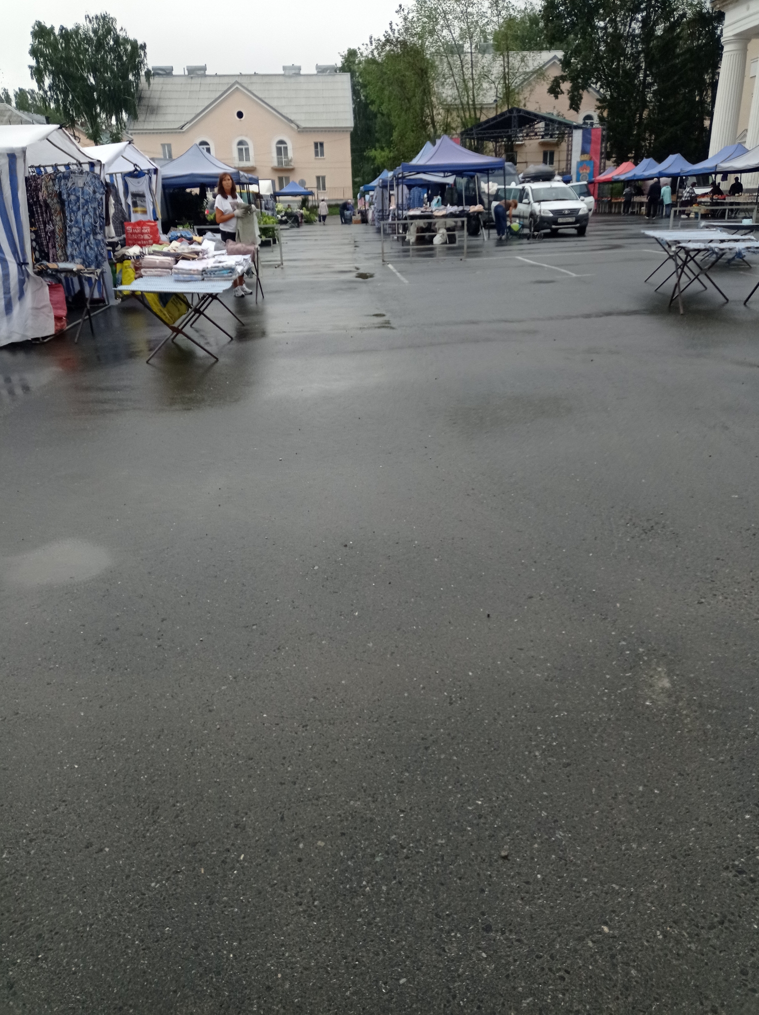 The image captures an outdoor market scene in Degtyarsk, Russia, likely on a cloudy or overcast day, as indicated by the wet asphalt and the diffused lighting. In the foreground, the wet asphalt surface is the dominant feature, reflecting the surroundings. To the left, a market stall with a white and blue striped awning displays various clothing items on racks. A woman in a white top and dark skirt is attending to a table laden with folded fabrics or clothing. In the middle ground, several other market stalls with blue and purple awnings are visible. A white car with its doors open is parked near one of these stalls, and a person is attending to it. Further back, more market stalls extend into the distance, with people browsing and interacting. The background features a light-colored building with arched windows and balconies, and behind it, trees and other structures. Russian flags are prominently displayed on a stage-like structure in the middle of the market. The overall impression is that of a bustling but somewhat subdued market environment, possibly impacted by the weather.