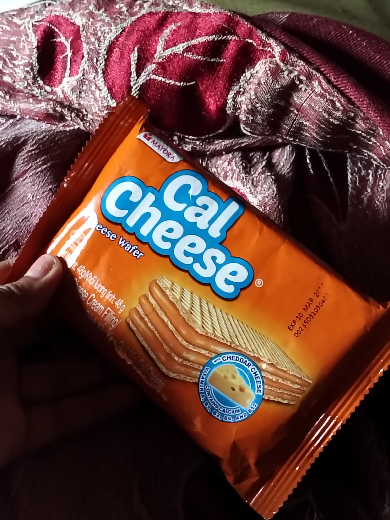 The image shows a person's hand holding a package of "Cal Cheese" wafers. The package is orange with blue and white lettering. The wafers themselves are visible through the packaging, showing stacked layers of wafer with a creamy filling. A circular emblem on the package indicates that it contains cheddar cheese and vitamins. Text on the package includes "MAYORA," "Cal Cheese," "Cheese Wafer," "48g," and an expiration date of "EXP:30 MAP 2007." The background is a textured, deep red fabric with intricate patterns. The scene appears to be an indoor setting, possibly a home or a store, given the casual way the product is held and the texture of the background. The lighting is warm, suggesting it could be daytime or indoor lighting. The overall impression is a close-up shot of a snack product being presented or examined.