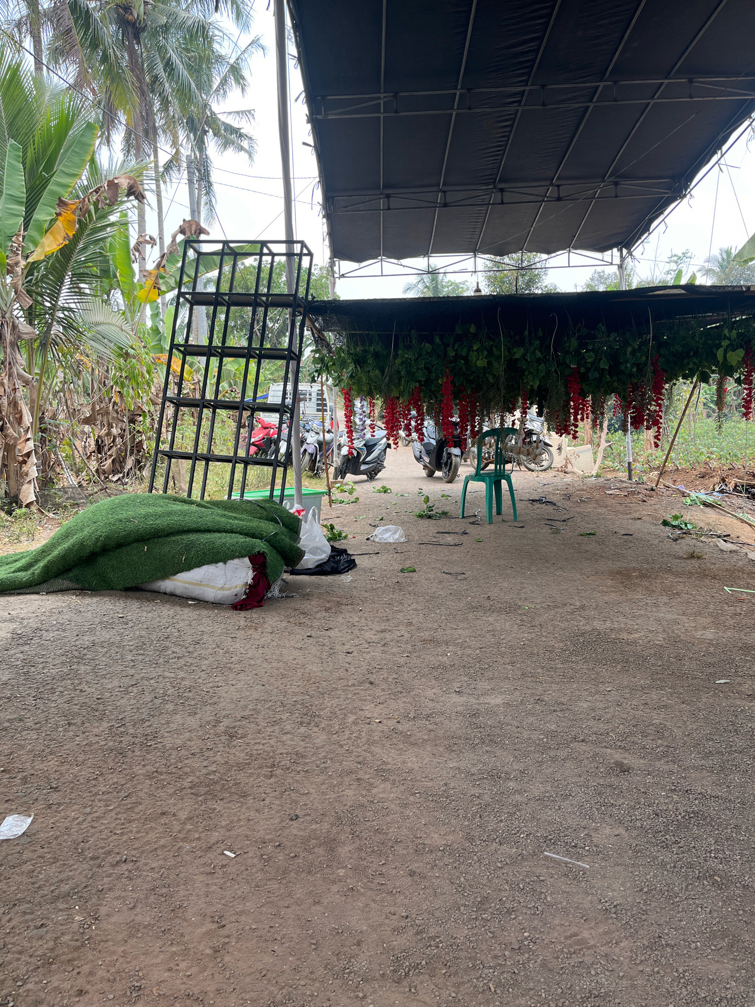 The image depicts an outdoor area, likely a market or event setup space, in Kebumen, Indonesia. A large roll of artificial turf is laid out on the ground to the left, partially covered by a red cloth. Behind it, a black metal shelving unit stands upright. Several motorcycles are parked in the background, some under a black canopy adorned with hanging red flowers and green foliage. A green plastic chair is placed in the center of the open space. The ground is unpaved, composed of dirt and gravel. In the distance, palm trees and other vegetation are visible. The overall scene suggests a temporary setup, possibly for an event or sale.