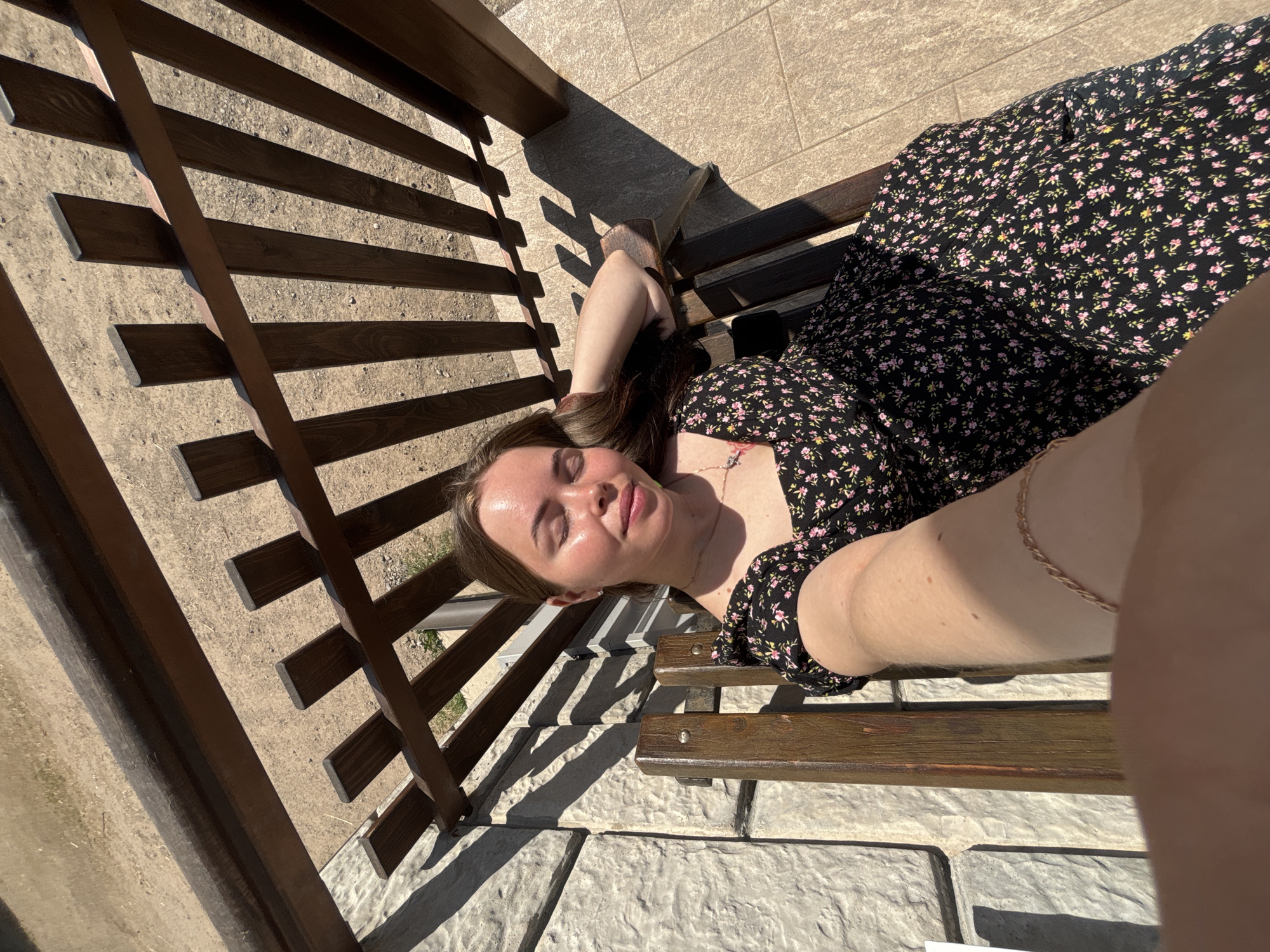 A young woman is lying on a wooden bench, seemingly outdoors, possibly in Русский Вожой, Russia. She is wearing a black dress with small pink and yellow flowers. The bench casts shadows on a light grey tiled surface. The surrounding environment includes wooden structures with lattice-like designs.