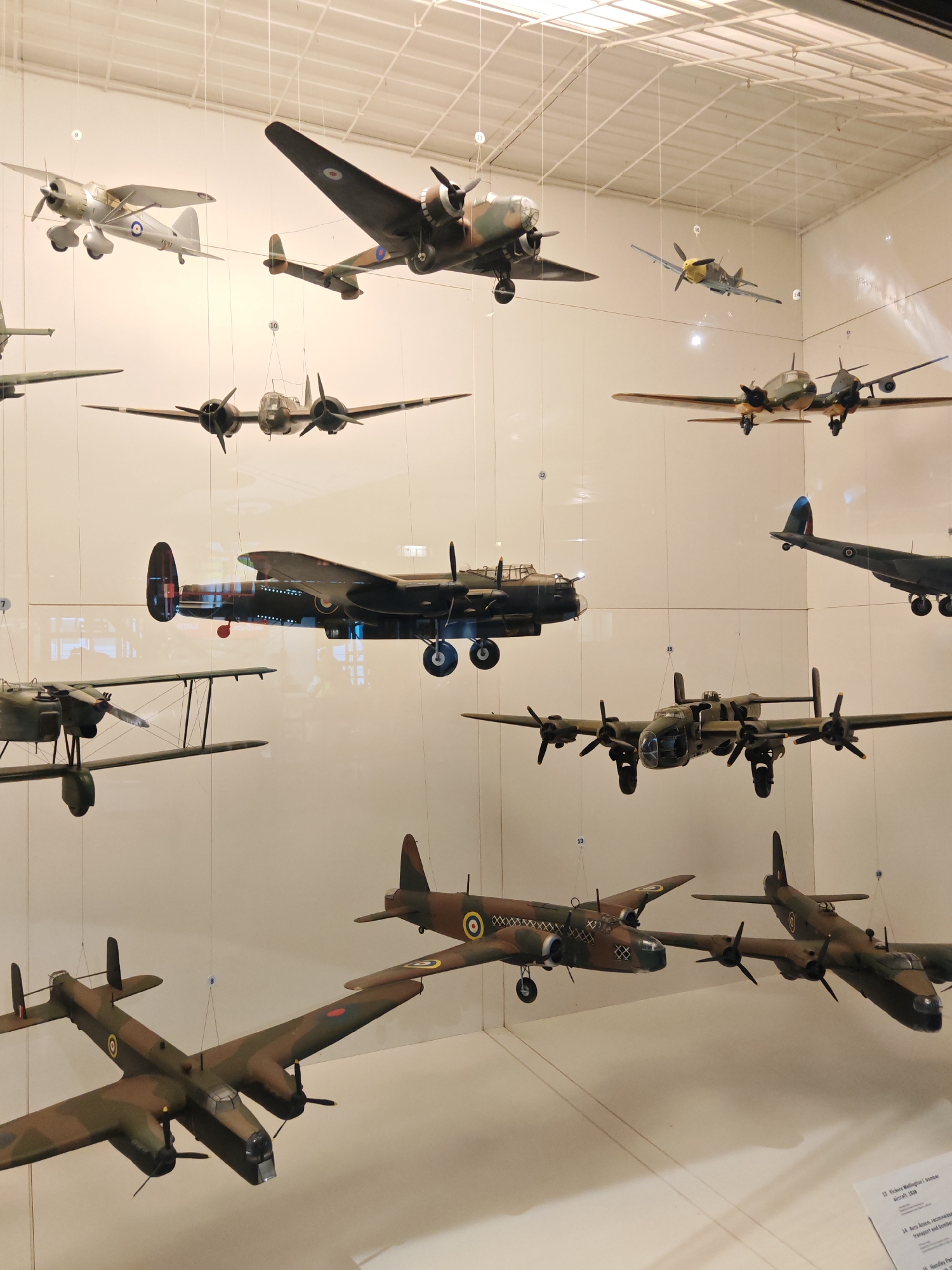 This image shows a collection of model airplanes on display, likely in a museum exhibit. The models are suspended from the ceiling by wires. The models feature a variety of World War II aircraft. There are several biplanes and multi-engine bomber models. The setting appears to be indoors, with a white wall as the backdrop. The text on the bottom right corner mentions "Vickers Wellington," which suggests that the image may be from the Royal Air Force Museum located in the city of London, United Kingdom.