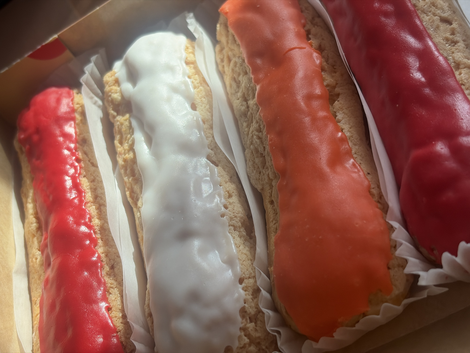 The image shows a box of four éclairs, each with a different colored glaze: red, white, orange, and red. They are arranged side-by-side in the box, separated by white paper liners. The éclairs appear to be freshly made and are the primary subject of the photo. There is no visible indication of the location being Saint Petersburg, Russia, no people, and no specific actions or interactions are apparent within the scene. The environment is simply the interior of the box with the éclairs.