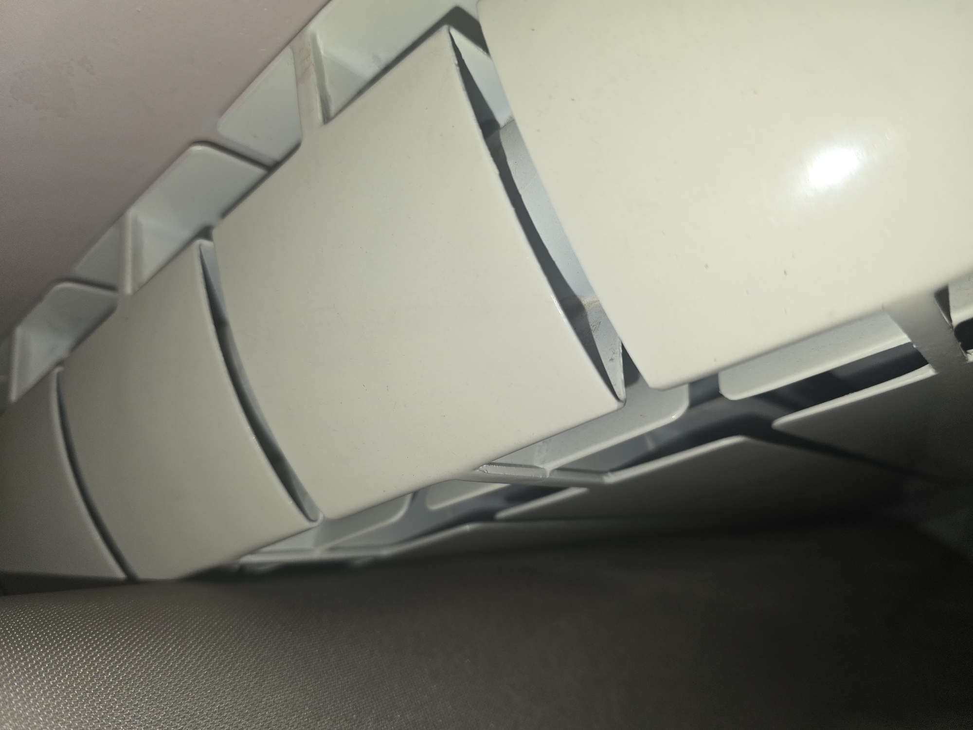 The image shows a close-up, angled view of a white radiator. The radiator has a segmented design, with each segment appearing to be a curved, elongated shape. The segments are arranged vertically and appear to be made of metal. To the left of the radiator, a textured gray surface, possibly a curtain or fabric covering, is visible. The background to the right of the radiator is a light pinkish-purple wall. The lighting suggests an indoor environment, and there are no people or any other identifying objects visible. The scene is captured from a slightly elevated perspective, looking down and to the side. The location in Ulan-Ude, Russia cannot be determined from the visual information provided in the image.