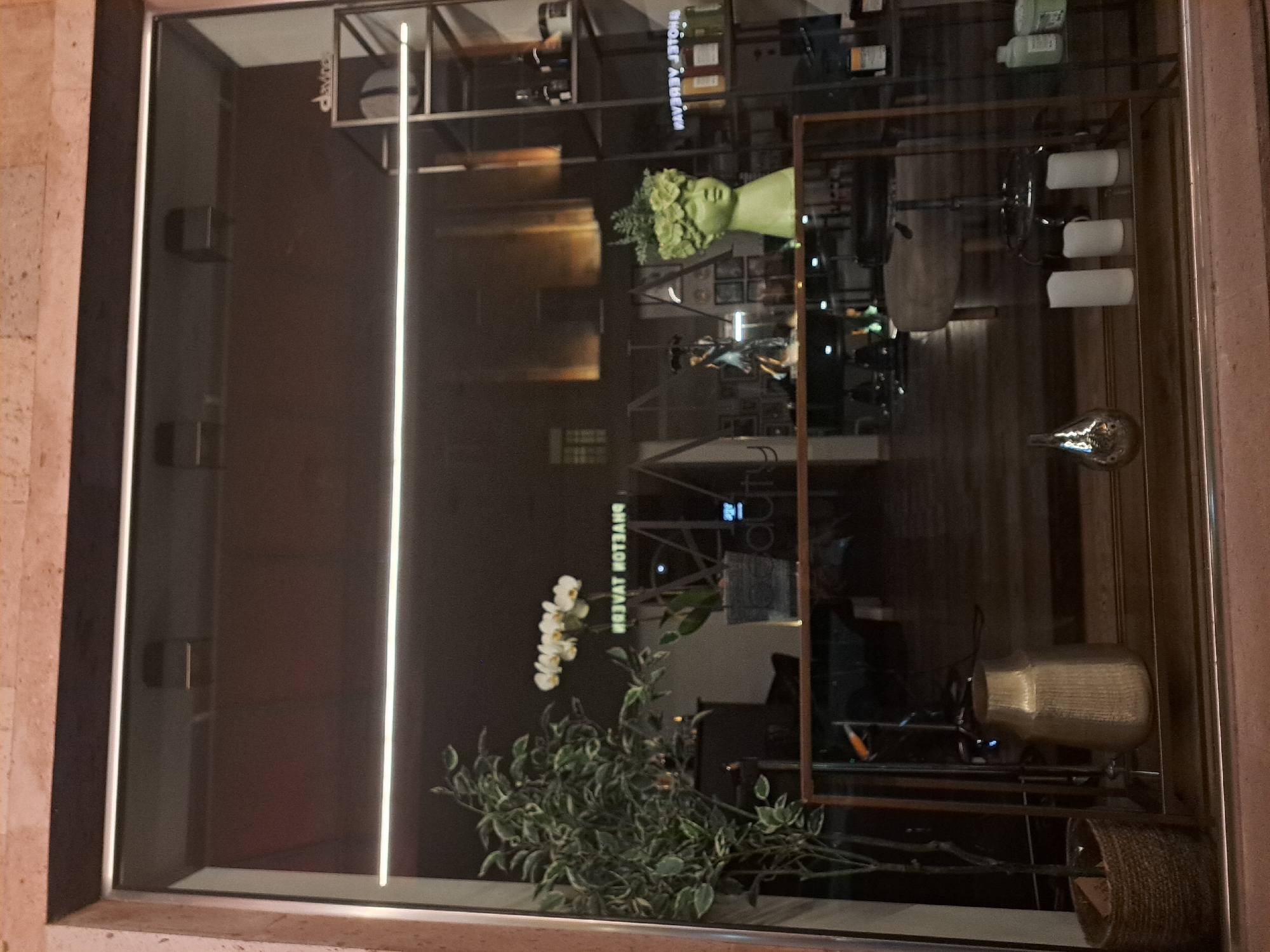 A window display in Yerevan, Armenia, features a layered arrangement of objects and reflections. A glass case contains shelves displaying bottles, a head-shaped bust with greenery, and other decorative items, including vases and rolled towels. A plant with white flowers and green leaves sits at the base of the display. Reflected in the glass is a blurred streetscape, with building facades visible in the distance. The window frame is dark and contrasts with the lighter exterior wall.