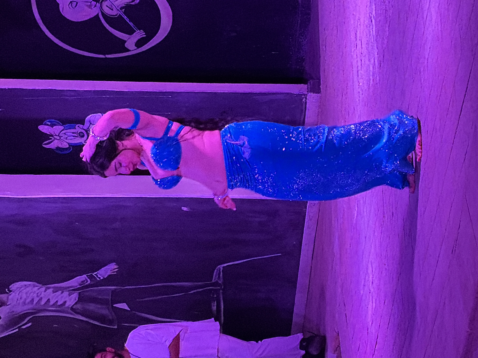 A woman in a blue sequined belly dancing costume is performing on a stage with a mural in Sharm El Sheikh, Egypt. The mural features cartoon-like drawings of characters. A man in a white shirt and dark pants is visible at the bottom left corner of the image. The stage floor is a light-colored wood. The lighting in the scene is predominantly purple.