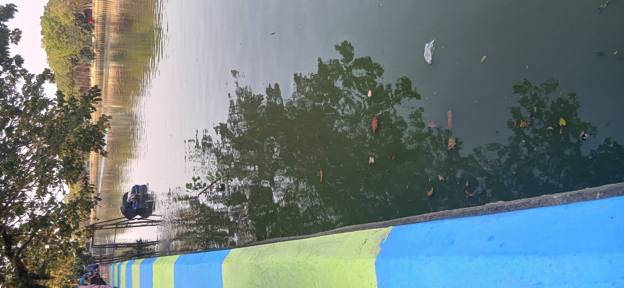 The image depicts a section of the Mahakam River in Samarinda, Indonesia, likely near a public recreational area. In the foreground, a concrete embankment, painted with horizontal blue and green stripes, borders the river. The river water is murky and reflects the surrounding trees. Scattered on the surface of the water are fallen leaves and some debris, including a white plastic bag and what appears to be a small, dark object partially submerged. In the mid-ground, a small blue boat with a canopy is tied to wooden posts. A few individuals are visible sitting along the embankment on the left side of the image, partially obscured by tree branches. The background shows a line of trees and vegetation along the riverbank, with the hazy sky visible above. The overall impression is of a slightly neglected urban riverfront environment.