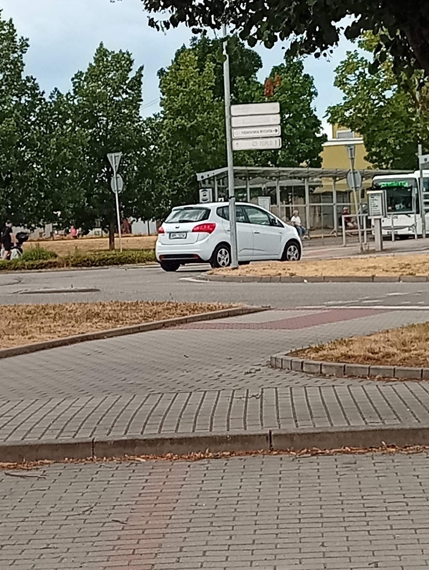 The image depicts a street scene in Tišnov, Czechia, featuring a white car parked on the right side of a road. Behind the car, a bus stop shelter with people waiting is visible. Trees and foliage frame the upper portion of the image, suggesting an outdoor setting in a town or city. The ground is paved with interlocking bricks, and dry grass lines the side of the road. A traffic sign with directional arrows and text, likely indicating local directions, is also present. The sky is overcast.