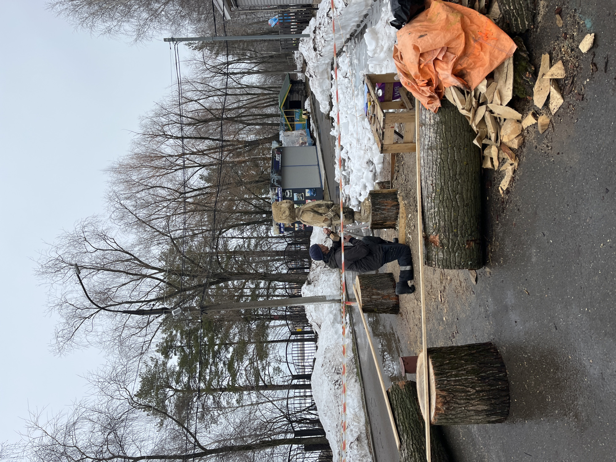 The image depicts an outdoor scene, likely in the city of Chaikovsky, Russia, given the presence of what appears to be a temporary market stall and surrounding urban infrastructure. A man dressed in dark clothing and a hat is working on a log, possibly carving or shaping it. He is positioned along a row of logs, presumably for his work. Wood chips are scattered around the log being worked on.
The scene is situated next to a street with snow piled on one side, separated by an orange-and-white striped barrier tape. Behind the barrier, there are trees without leaves and what appears to be a small commercial structure. Utility poles with wires are visible in the background against an overcast sky. The ground is wet, possibly due to melting snow.