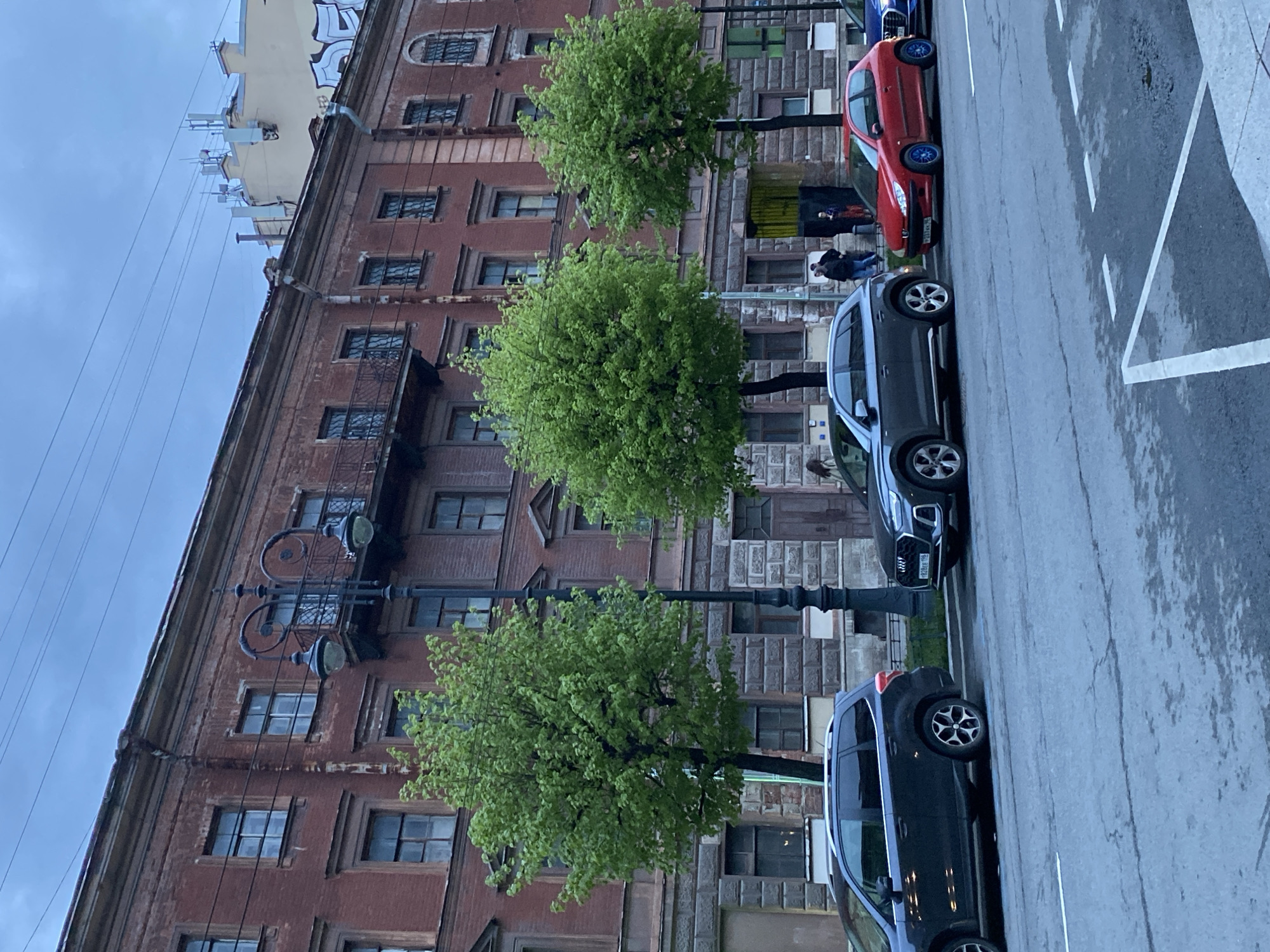 An eye-level, urban street scene is taken on a cloudy day with a red brick building situated on the left side of the frame. The street is lined with three trees, each with a round, dense canopy of green leaves, positioned in a row along the sidewalk. A black lamppost is placed between the lower and middle trees. The street is adjacent to the brick building. Parked cars are positioned along the street on the right side. One is a red passenger car, and another is a gray Audi SUV. A black car is in the foreground. Two individuals are walking on the sidewalk near the middle tree, behind the red car. The area is identified as Saint Petersburg, Russia, due to the Cyrillic characters on the license plate of the gray Audi. The sidewalk is marked with white painted lines.