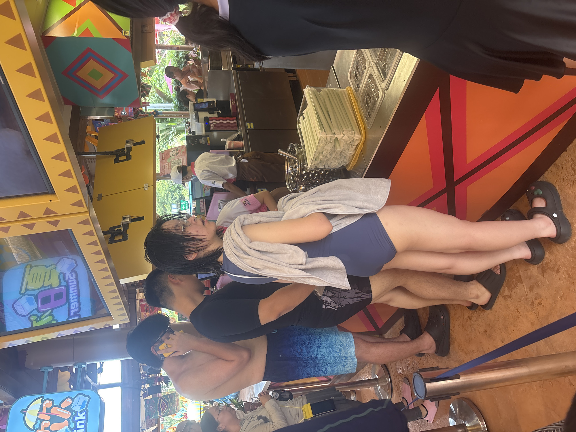 In Panyu District, China, a group of people are queuing at a food or drink stand, likely within a theme park or resort, given the colorful decorations and casual attire. The individuals are dressed in swimwear and light clothing, suggesting a warm climate. The primary focus is on the people at the counter, where drinks or snacks are being prepared or served. One person in the foreground is wearing a blue swimsuit and has a towel draped over their shoulder. To their left, a man in black shorts and a black t-shirt is visible. Further left, a man in blue and white patterned swim shorts is talking on his phone. In the background, park staff are working behind the counter, serving customers. The overall atmosphere appears to be relaxed and recreational.