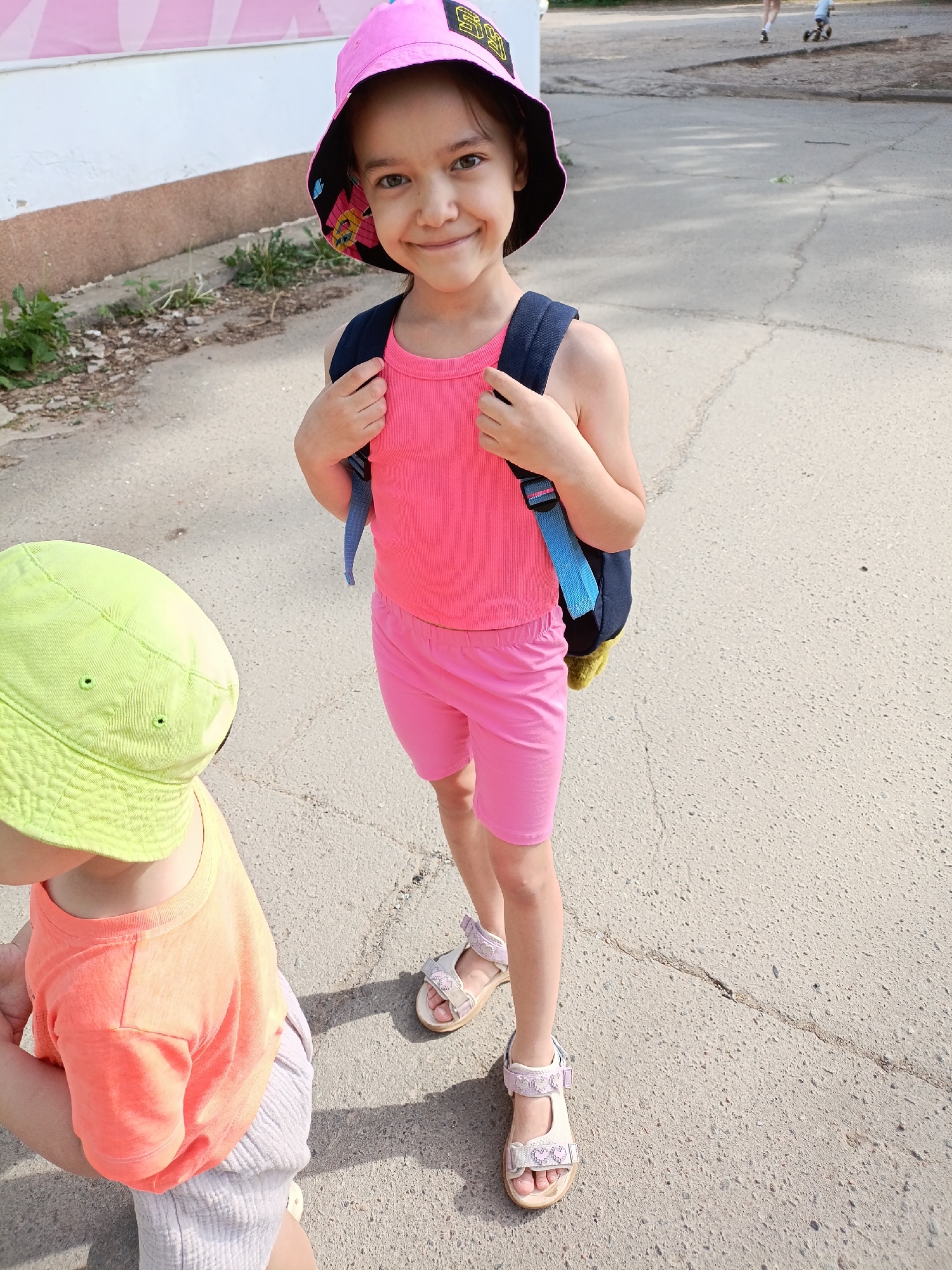 The image shows a young girl standing on a paved surface. She has brown hair and eyes, and is wearing a pink bucket hat, a pink tank top, pink shorts, and sandals. She is also wearing a blue and yellow backpack. To her left is a younger child wearing a green hat and orange t-shirt. The background shows a building with a pink sign and another pair of children farther down the road. Based on prior knowledge, this image may have been taken in the city of Chaikovsky, Russia, but there is no detail in the image to verify the location.