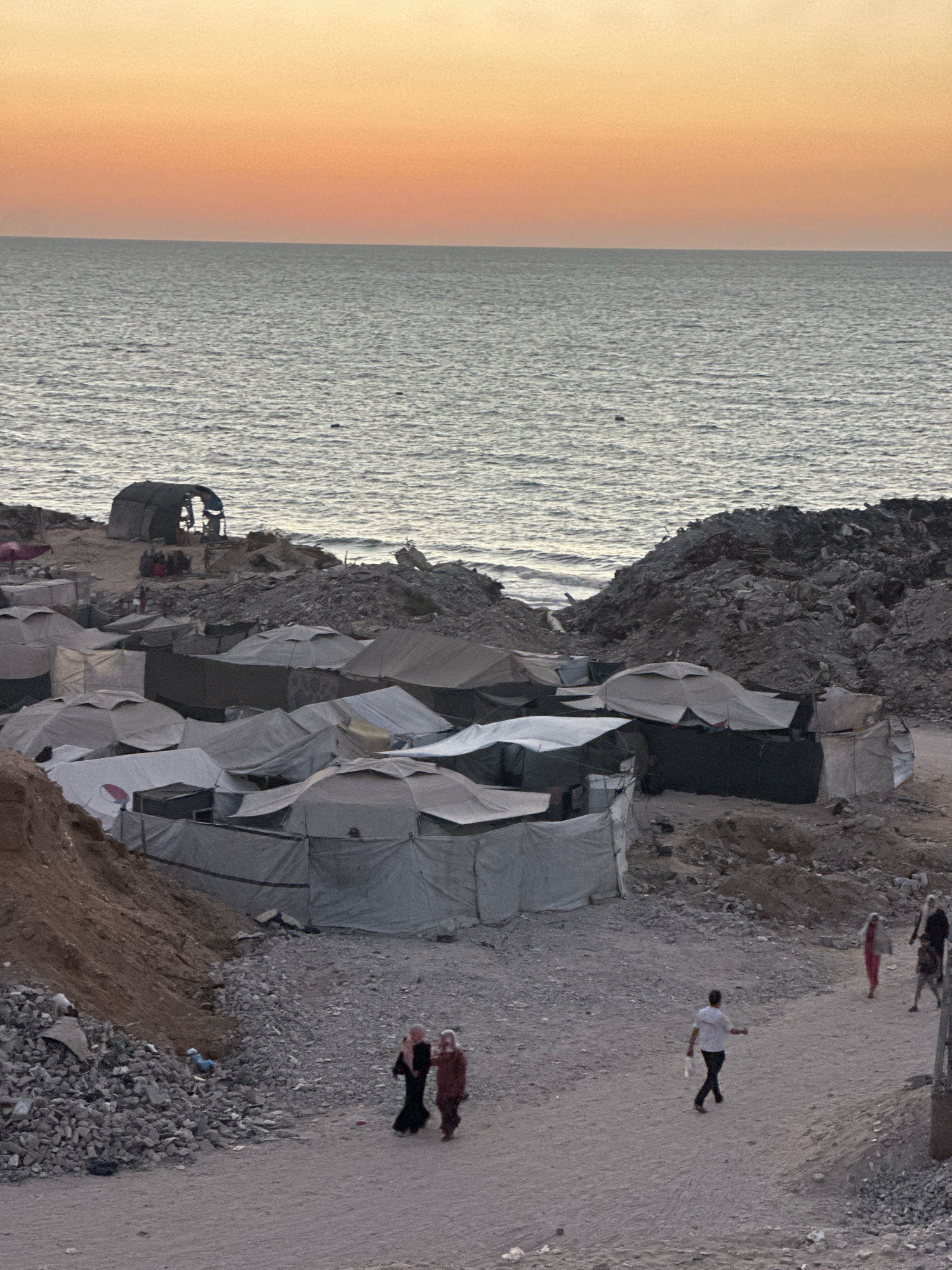 This image depicts a coastal view of the city of Gaza, Palestinian Territories, at sunset. The sky transitions from a soft orange at the horizon to a pale yellow, with the calm, gray sea occupying the upper half of the frame.
In the foreground, a densely packed tent city is visible, suggesting a displacement or refugee camp. These temporary structures are made of white and gray fabric, with some appearing to be older and in disrepair. The tents are clustered closely together, interspersed with piles of rubble and dirt.
Several people are seen walking along a dirt path that winds through the camp. Two individuals in the lower center are dressed in traditional clothing, with headscarves. Further up the path, a man in a white shirt and dark pants is walking, carrying a white bag. On the right side of the image, two women and a child are walking away from the camera.
In the background, closer to the sea, a larger, more industrial-looking tent or shelter is partially collapsed. This area is also characterized by significant amounts of debris and rough terrain. The overall impression is one of a challenging living environment, with the sea providing a stark backdrop to the makeshift dwellings.