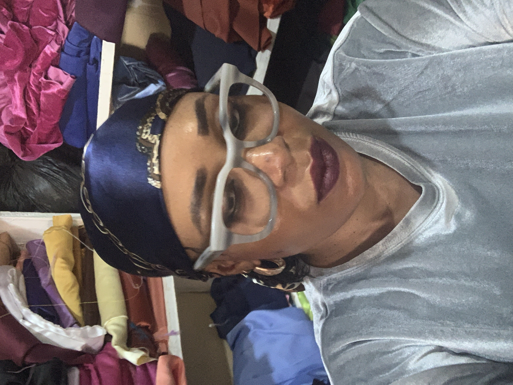 A woman with dark hair and skin is wearing a blue headscarf with gold accents and silver-framed eyeglasses. She has a light grey or silver shirt. Her lips are painted a dark plum color. She appears to be indoors, surrounded by draped fabrics in various colors. Based on the description of the location, this photo would have been taken in Sabon Tasha, Nigeria.