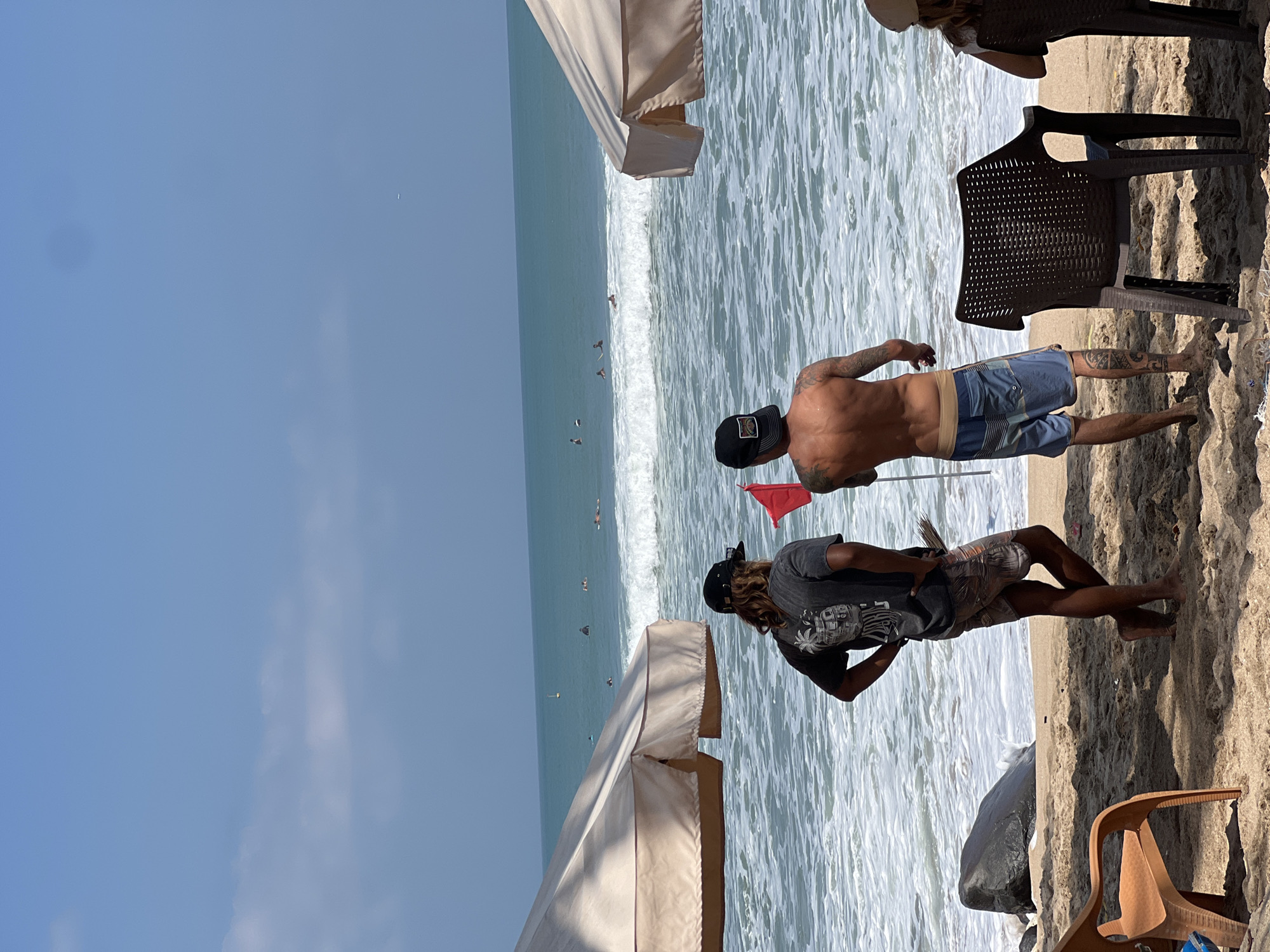 Two men stand on a sandy beach, facing the ocean. The man on the left has long, wavy hair and wears a dark t-shirt and patterned shorts. The man on the right is shirtless and wears blue swim trunks. He holds a red flag. In the background, several surfers are in the water, with waves breaking. Two large, cream-colored umbrellas provide shade on the beach. The scene appears to be in Kuta, Indonesia, a popular beach destination known for its surf.
