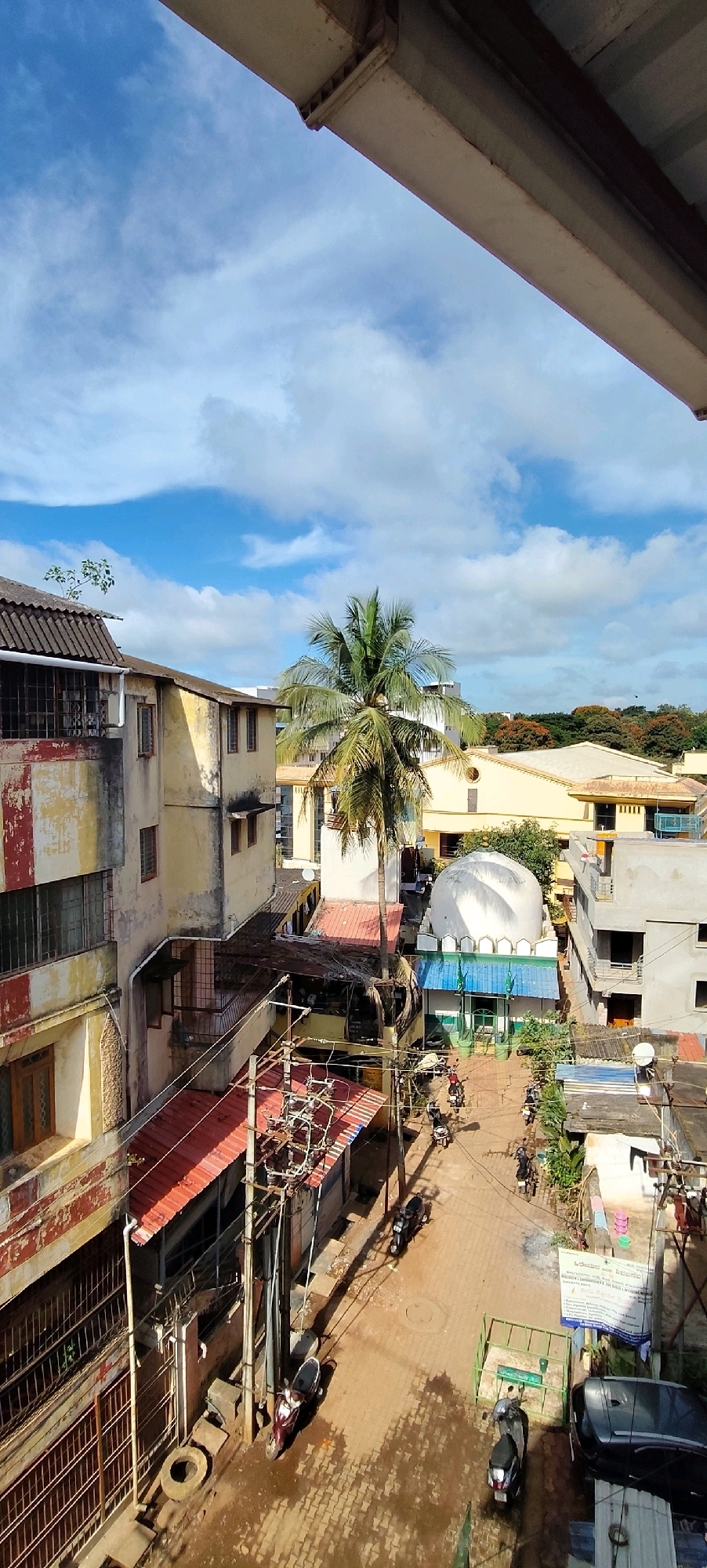 The image captures an aerial view of a street in Belagavi, India, under a partly cloudy sky. A prominent palm tree stands near the center of the frame, beside a white dome-shaped structure with green accents and blue roofing, resembling a mosque or shrine. Several multi-story buildings with weathered facades in yellow, red, and white line the street. Electrical poles with numerous wires are visible, indicating urban infrastructure. The street itself is paved with tan interlocking bricks and appears wet in places, reflecting the sunlight. Several motorcycles and a car are parked or being ridden on the street, suggesting moderate activity. The overall scene depicts a dense urban environment with a mix of residential and religious buildings.