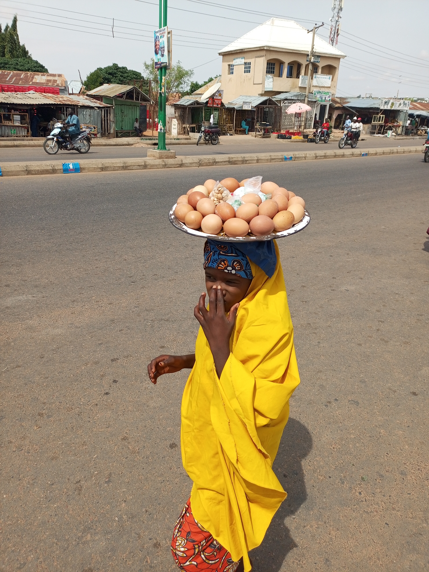 A young Black person wearing a yellow headscarf and yellow garment, with a colorful skirt, is walking on a paved road, carrying a large metal tray of eggs on their head. They are holding their right hand to their face. In the background, there are buildings, shops, a traffic light, and people on motorcycles. The scene appears to be a busy street in a city.