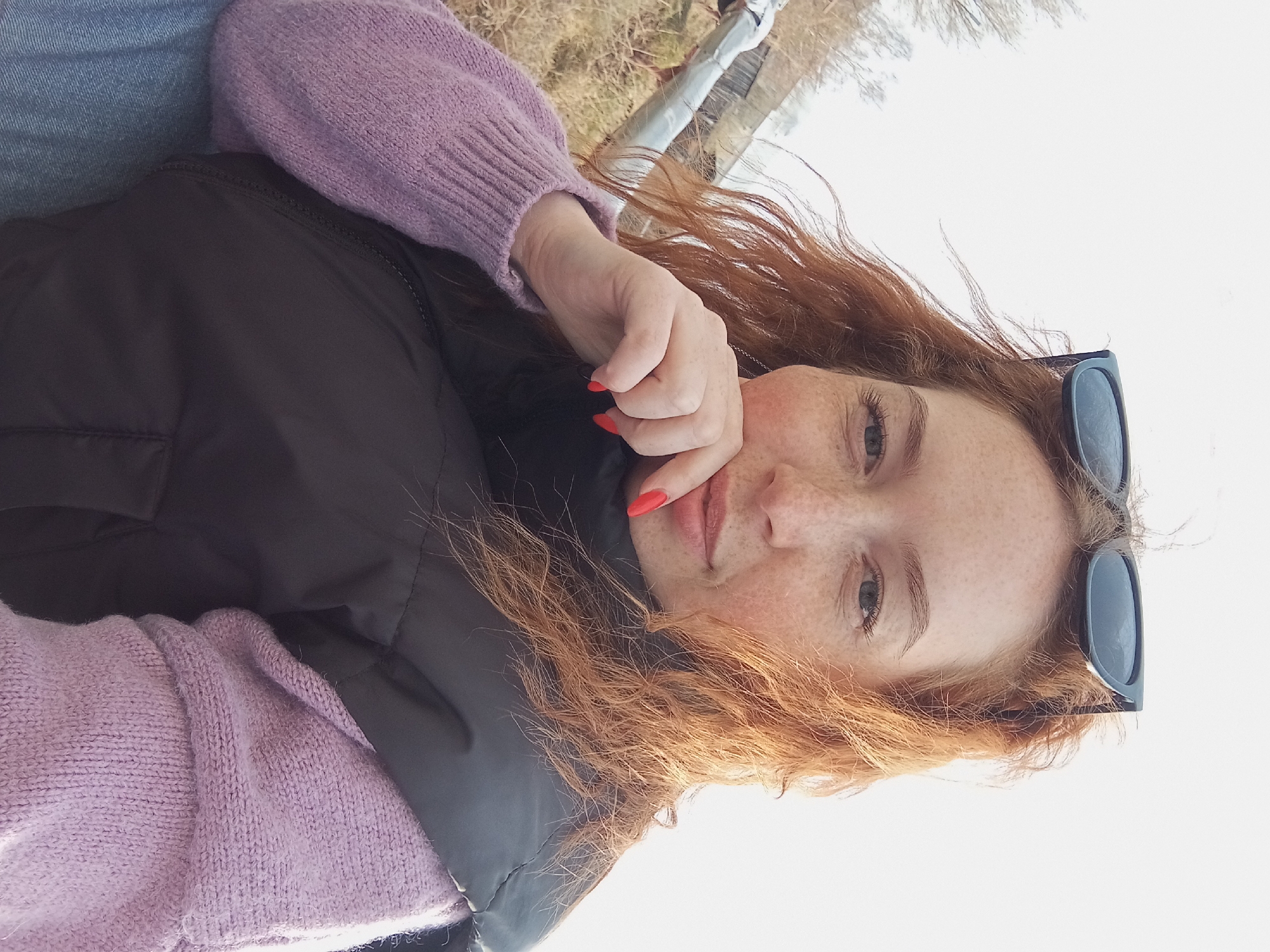A woman with red hair and freckles is wearing a lavender sweater and a black vest. Sunglasses are perched on top of her head. Her nails are painted red. The location is Yurgamysh, Russia. The woman is holding her hand to her face. The background appears to be trees and a white sky.