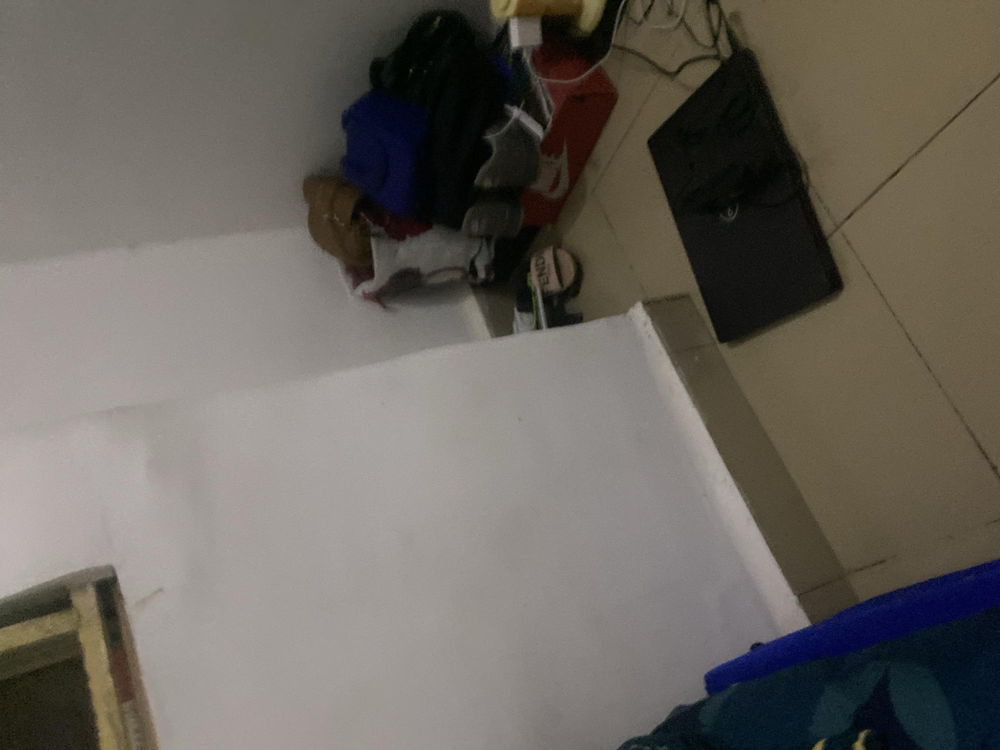 The image depicts a cluttered corner of a room, likely a bedroom or storage area, with items piled on the floor and against the walls. In the foreground, a dark-colored laptop is placed on a tiled floor. To the right of the laptop, various objects are visible, including a yellow cylindrical item that appears to be a lamp or speaker, connected to wires. Behind these items, a red shoebox is prominent, with shoes and bags stacked on top and around it. A pair of light pink sandals with "FENDI" written on them are visible on the floor near the wall. In the background, a white wall meets another wall that appears to be a lighter beige or cream color. A shelf with some items is partially visible in the upper left corner of the image. The overall impression is one of disarray, with personal belongings scattered in a living space. There are no people visible in the image. The location in Howayi, Nigeria, is not discernible from the visual information provided.