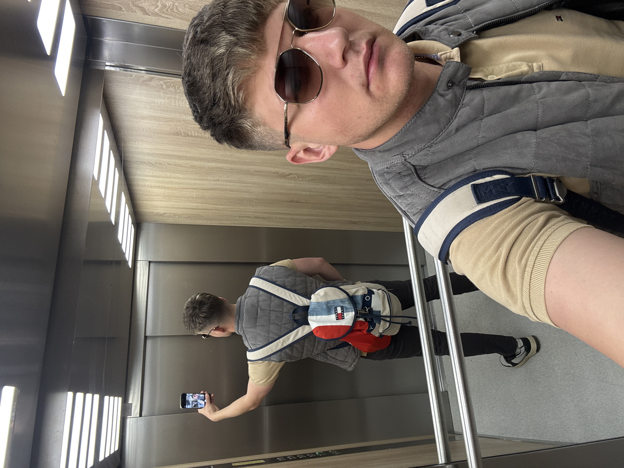 A man is taking a selfie in an elevator. The elevator has metallic walls and a wooden panel towards the top. The lighting consists of vertical LED strips. A second man is visible in the reflection on the back elevator wall, holding a cell phone and taking a picture. The first man is wearing sunglasses, a beige polo shirt, and a grey quilted vest. The second man is wearing a similar vest and a Tommy Hilfiger backpack. The floor appears to be grey. The location is Plovdiv, Bulgaria.