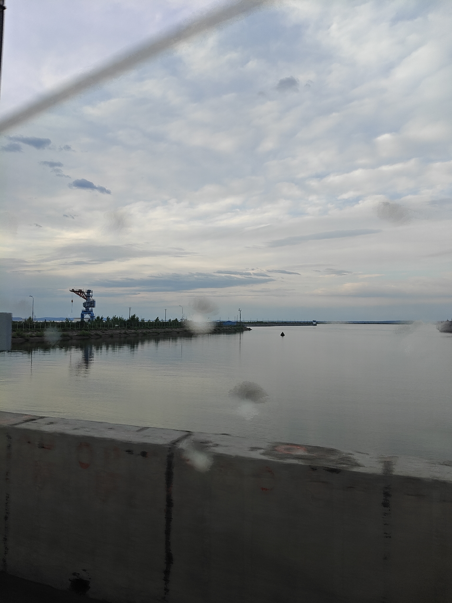 The image, likely taken from a moving vehicle, shows a cloudy sky and a body of water. A concrete barrier is visible in the foreground. On the far side of the water, there is a dock or port area with a crane structure. The water is calm, with reflections of the sky and the structures on the far bank.
Based on the location, it is likely the Kama River with the Port of Chaikovsky in Chaikovsky, Russia.