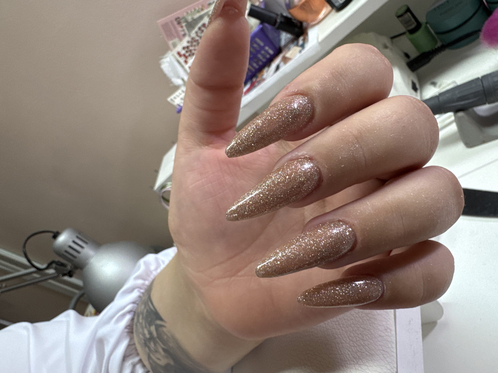The image shows a close-up of a person's hand with glittery, stiletto-shaped fingernails. The nails are a light brown or rose gold color with a high-gloss finish and fine glitter particles. The hand is resting on a white surface, possibly a table or a manicure station. In the background, out of focus, are various items that appear to be related to nail care or beauty services, including bottles, tubes, and what looks like a nail drill machine. There is also a silver desk lamp with a segmented neck. A tattoo is visible on the person's forearm. The environment suggests an indoor setting, likely a nail salon or a home beauty setup. There is no information available about the location in Ulyanovsk, Russia.