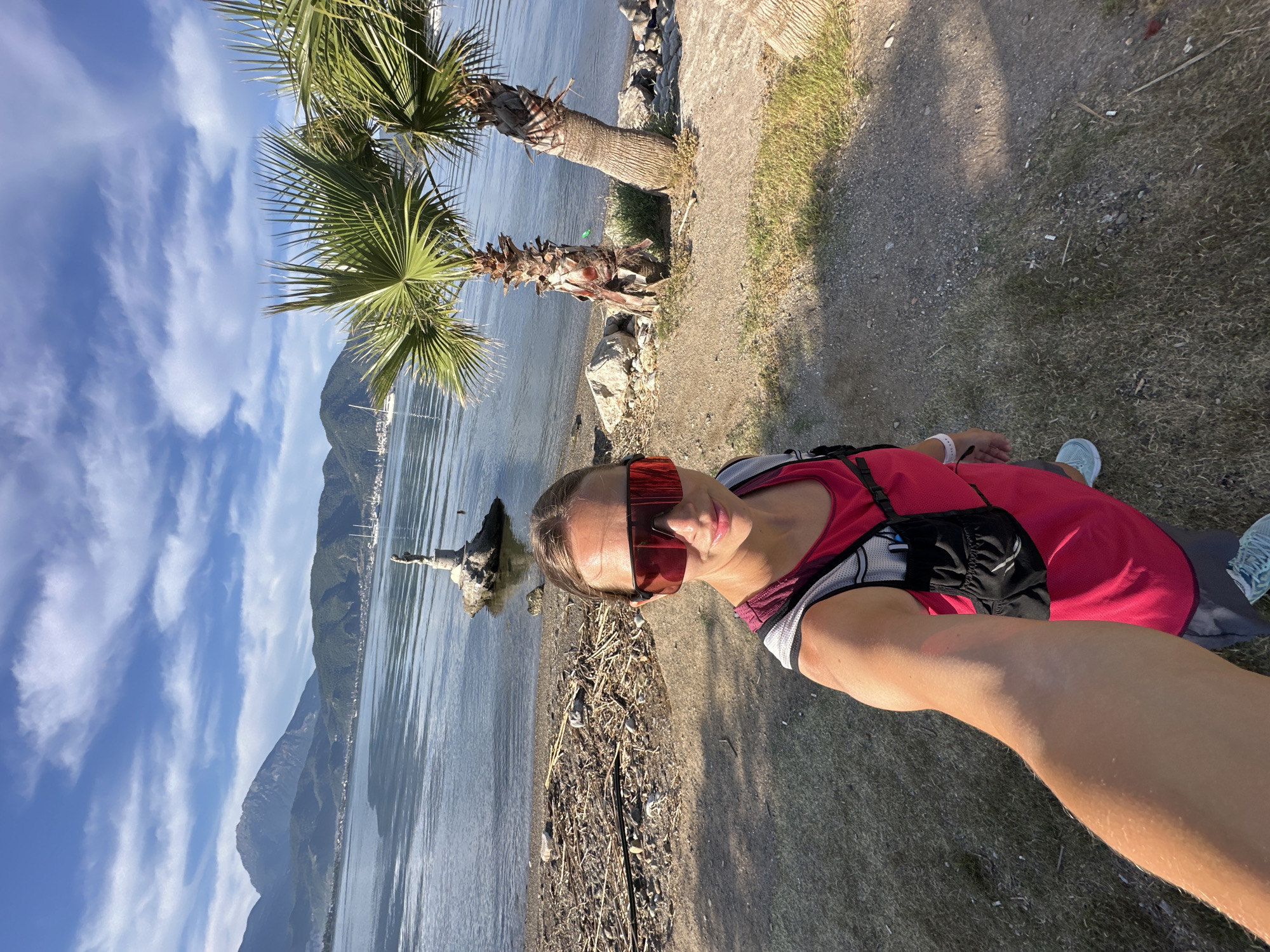 The image depicts a woman taking a selfie on a waterfront in Fethiye, Turkey. She is wearing red sports clothes and orange sunglasses, and she is on a path next to the water. Behind her are two palm trees, the coastline, mountains in the distance, a blue sky with white clouds, and the sea with a piece of driftwood. The ground is a mix of dirt, gravel, and grass.