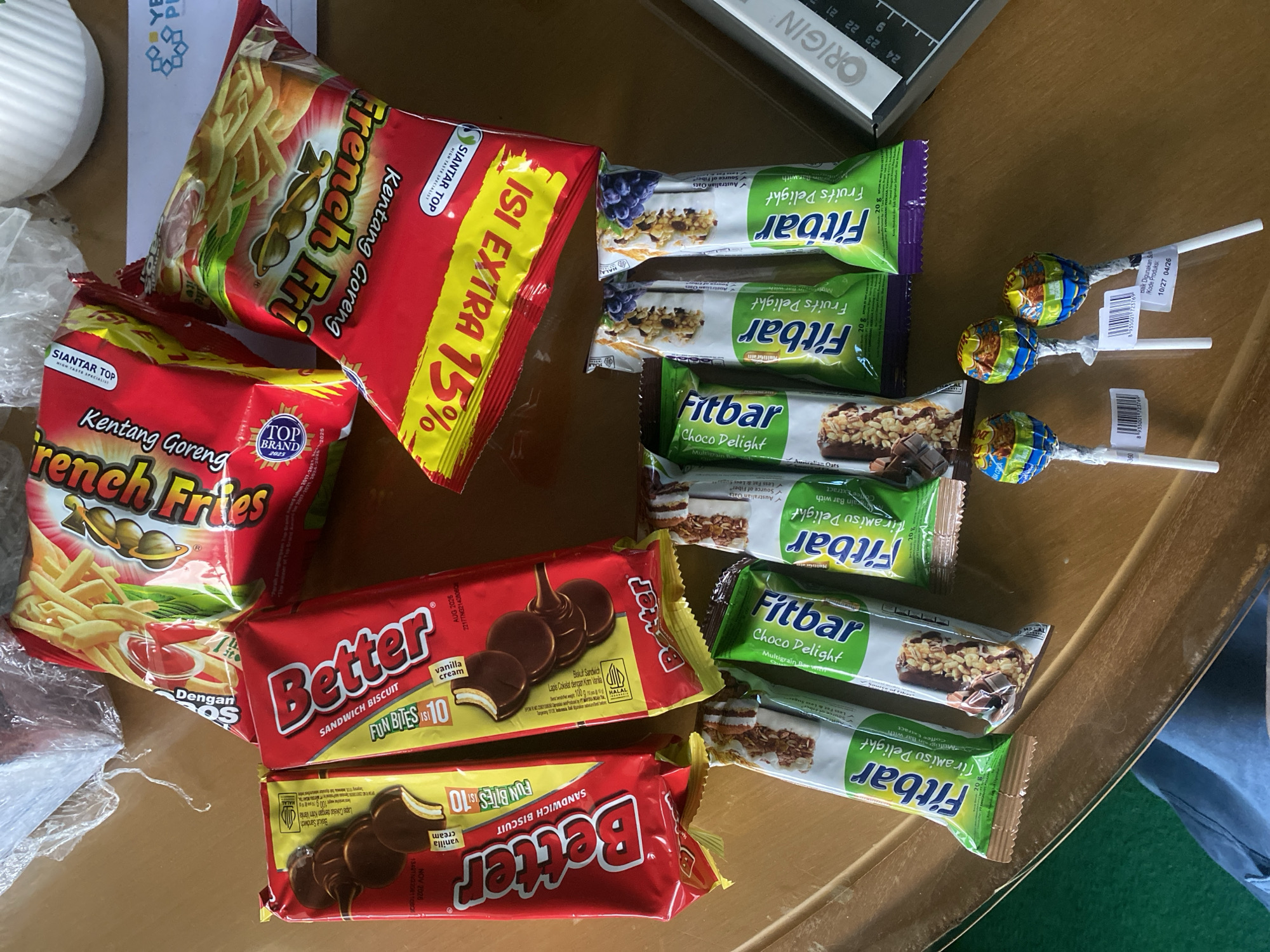 The image shows a collection of snacks and candies arranged on a wooden table. Among the items are several packages of "Better" sandwich biscuits, which are red and yellow with pictures of chocolate-covered biscuits. There are also packages of "Fitbar" snacks, in green wrappers, some labeled "Choco Delight" and others "Tiramisu Delight" and "Fruits Delight." Two red and yellow bags of "French Fries" potato snacks are also visible, featuring images of french fries and ketchup. Three colorful lollipops are positioned in the foreground. The background includes a white object and a piece of paper with text and a logo. The overall scene suggests a casual setting where snacks are displayed, possibly for consumption or sale.