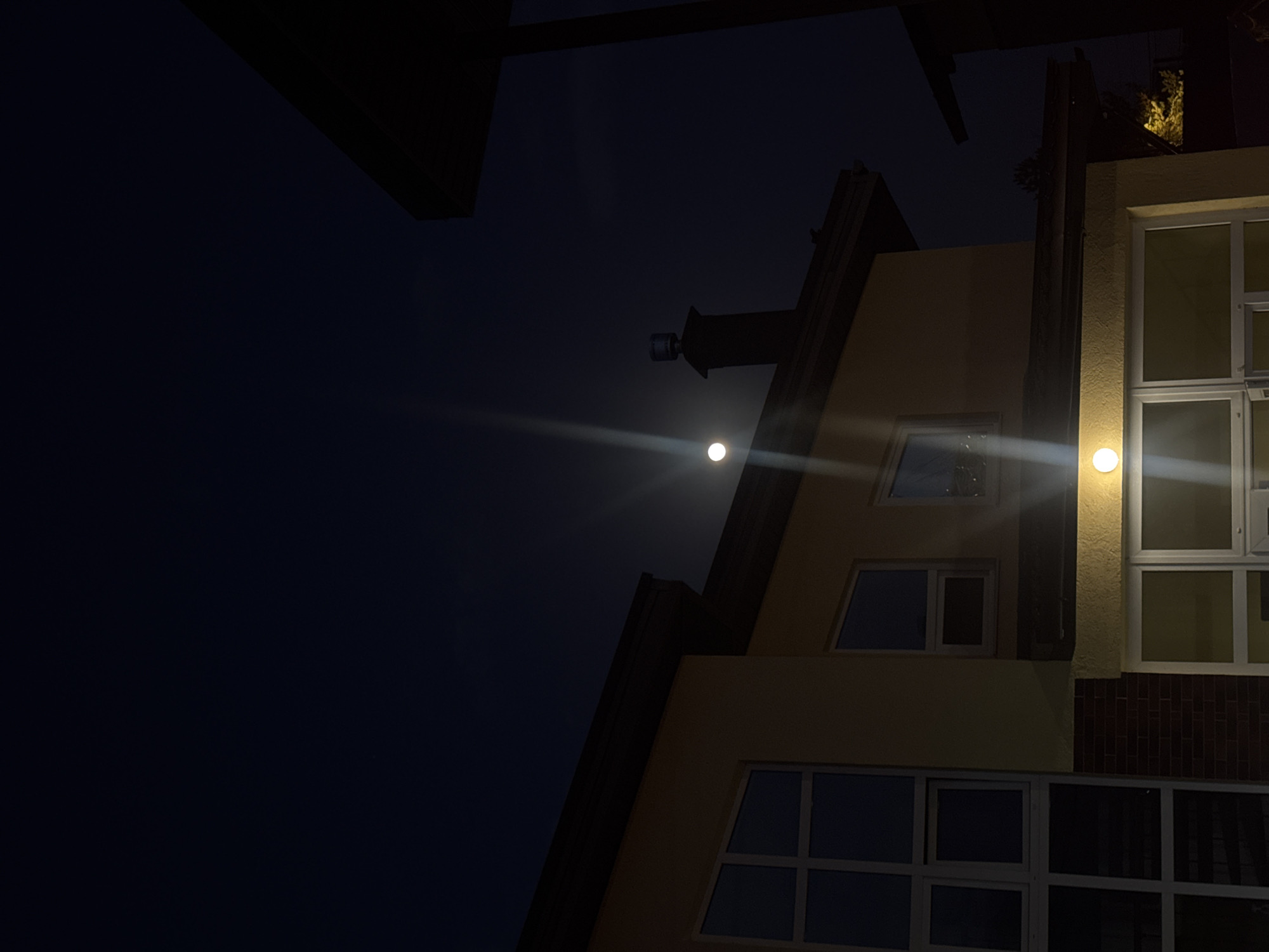 A low-angle shot captures a multi-story building in Ptichnoye, Russia, at night. The building's exterior is painted in shades of beige, and a section features red brick. Several windows are visible, reflecting the dark sky. A bright outdoor light fixture illuminates a portion of the facade. In the upper portion of the frame, the night sky is dark. A faint light source, potentially the moon, is visible in the distance, casting a streak of light across the sky. Overhanging architectural elements are seen in the extreme top, obscuring the scene.