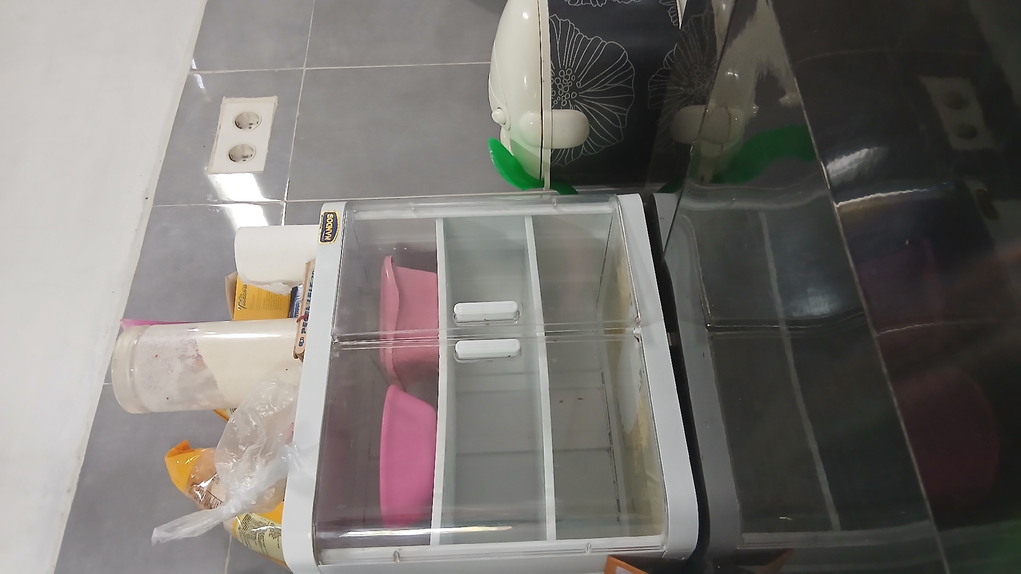 The image depicts a kitchen scene in Klambir Lima Kebun, Indonesia. In the foreground, a reflective black countertop takes up the bottom portion of the frame, showing reflections of items above. To the left, a clear plastic cabinet with white trim and doors sits on the countertop. The cabinet has two shelves. The top shelf holds two pink plastic bowls. The bottom shelf is empty and appears slightly dirty. The cabinet has a "NANDOS" logo on its top right corner. To the right of the cabinet is a black container with a white lid adorned with a floral pattern. A green object, possibly a handle or lid opener, is attached to the container's lid. In the background, a tiled wall is visible, with grey tiles forming a grid pattern. A double electrical outlet is mounted on the wall. On top of the plastic cabinet, various items are present, including a large plastic jar filled with a white powder, a yellow packet, and a clear plastic bag containing items. A roll of white paper is also visible. The overall impression is of a functional, albeit somewhat cluttered, kitchen workspace.