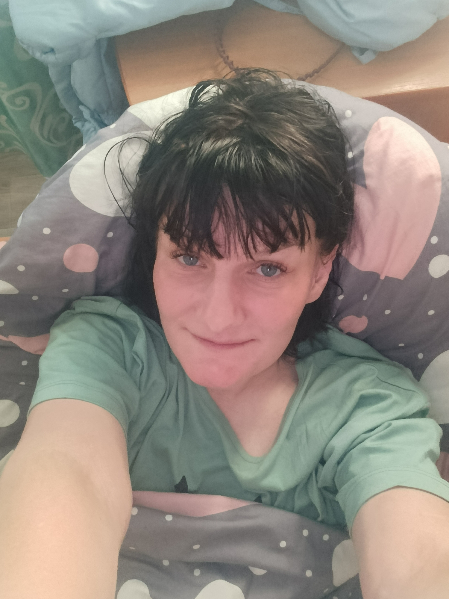 A person with dark hair, bangs, and blue eyes lies on a pillow with grey and white polka dots and pink heart-shaped designs. The person is wearing a light green t-shirt and appears to be taking a selfie. The background includes a wooden surface, a blue pillow, and a patterned green fabric. The location, Фоки, Russia, is not ascertainable from the image.